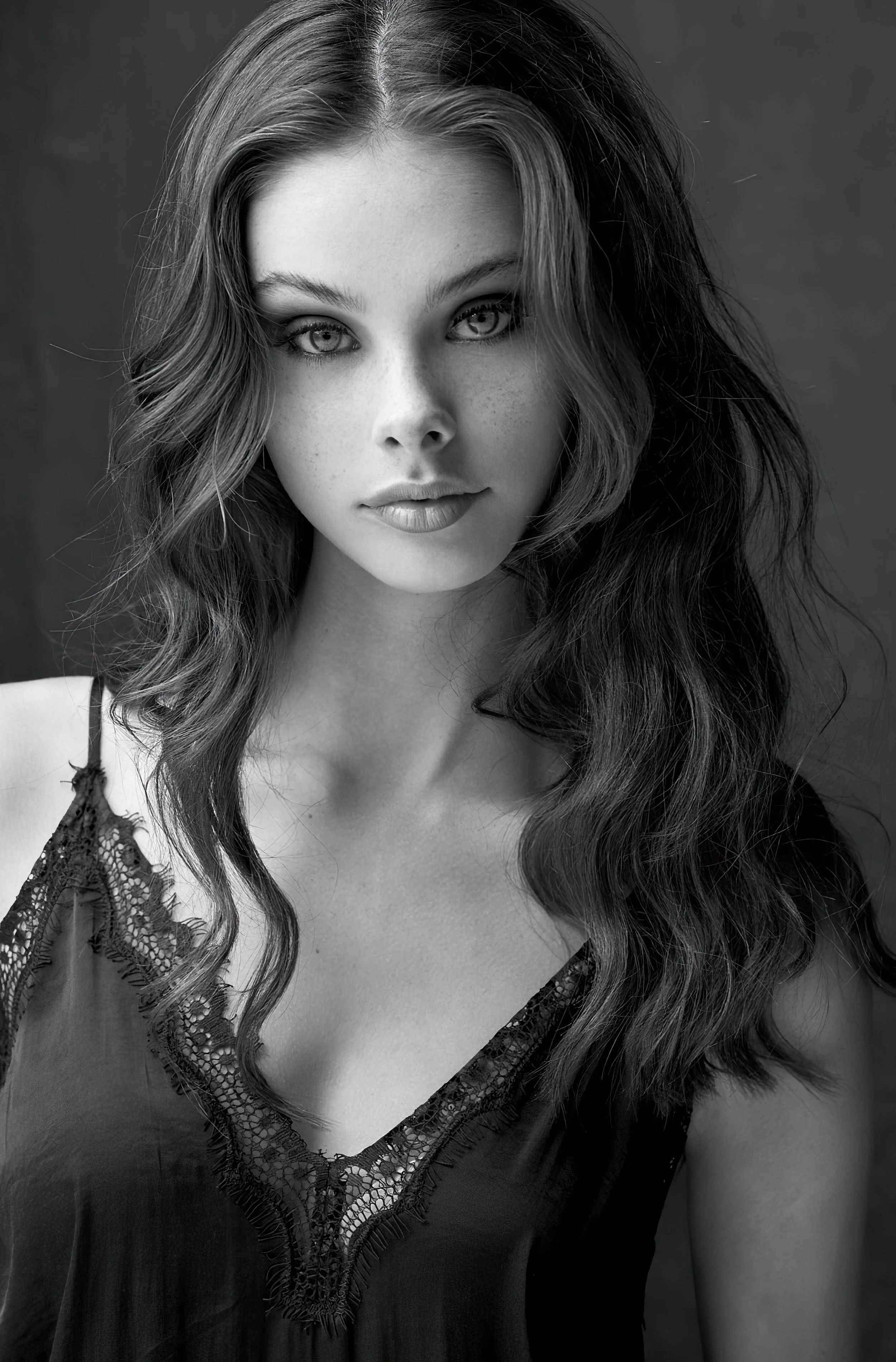 A black-and-white portrait of a young woman with wavy hair, wearing a lacy black dress, looking directly at the camera.