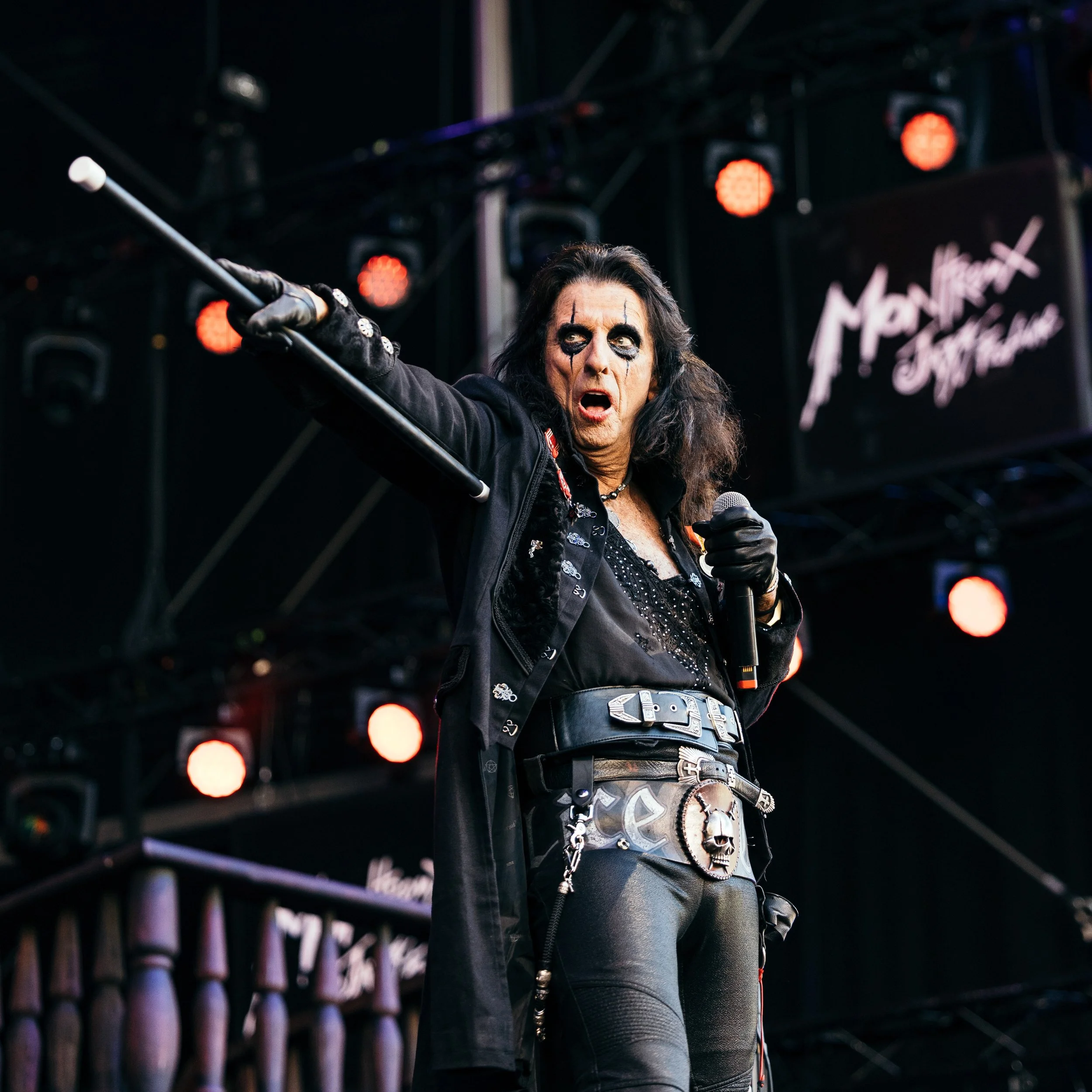 A rock singer with dark hair, black makeup, and a black outfit holding a microphone and pointing outward on stage during a performance.
