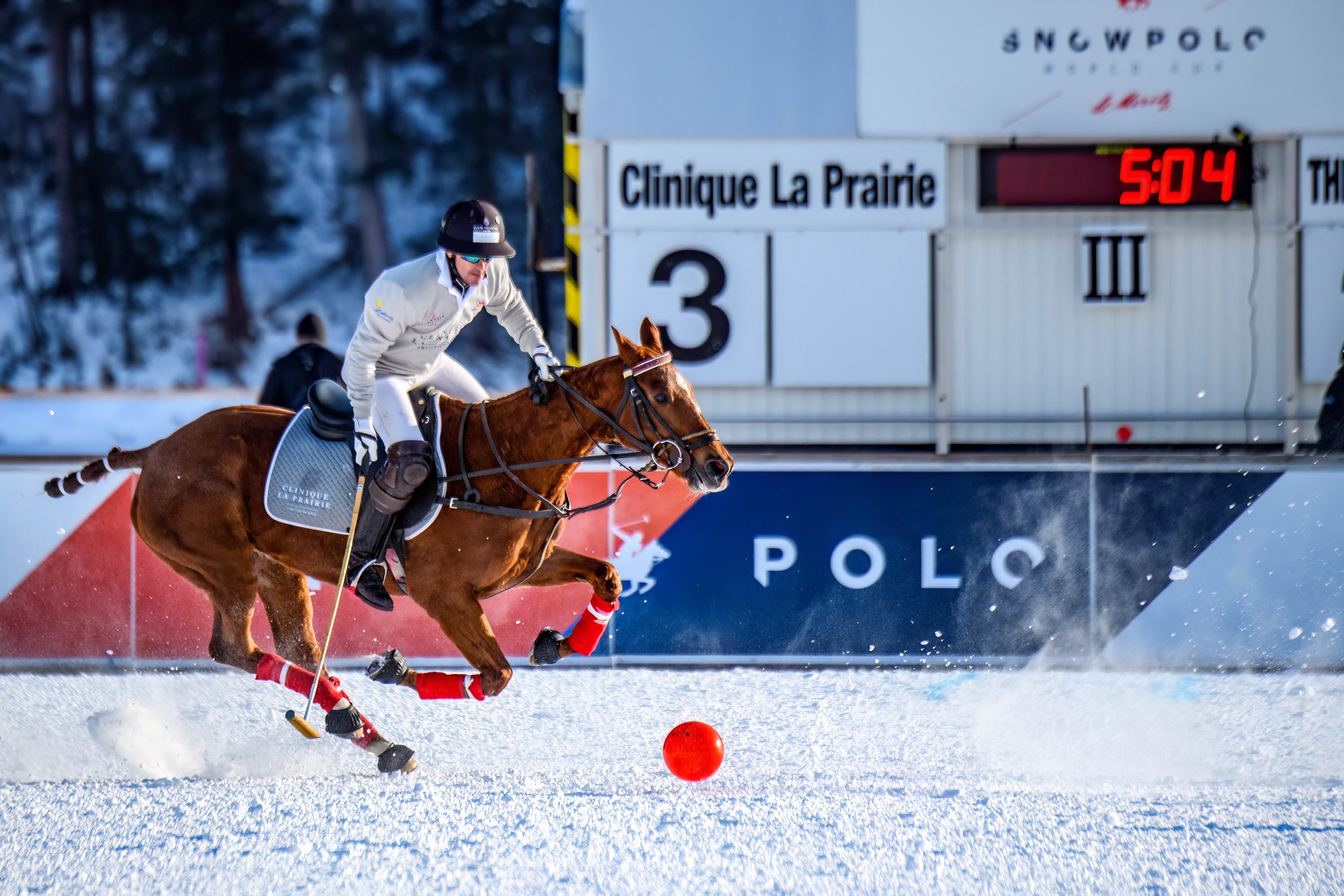 A polo player riding a horse on snow during a game, with a scoreboard in the background displaying 5:04 and the words Clinique La Prairie.