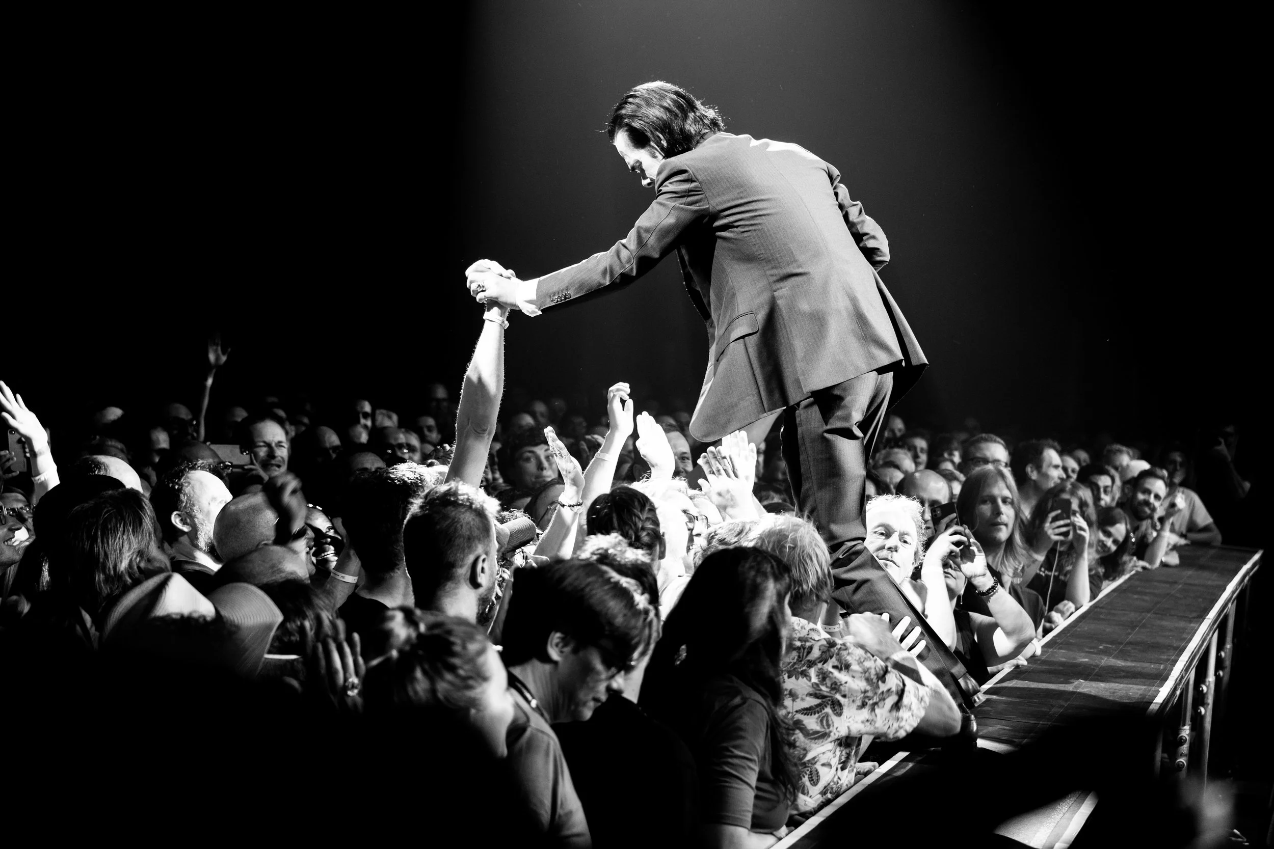 A man in a suit bends over a crowd, shaking hands and engaging with them during a performance or event, black and white photo.