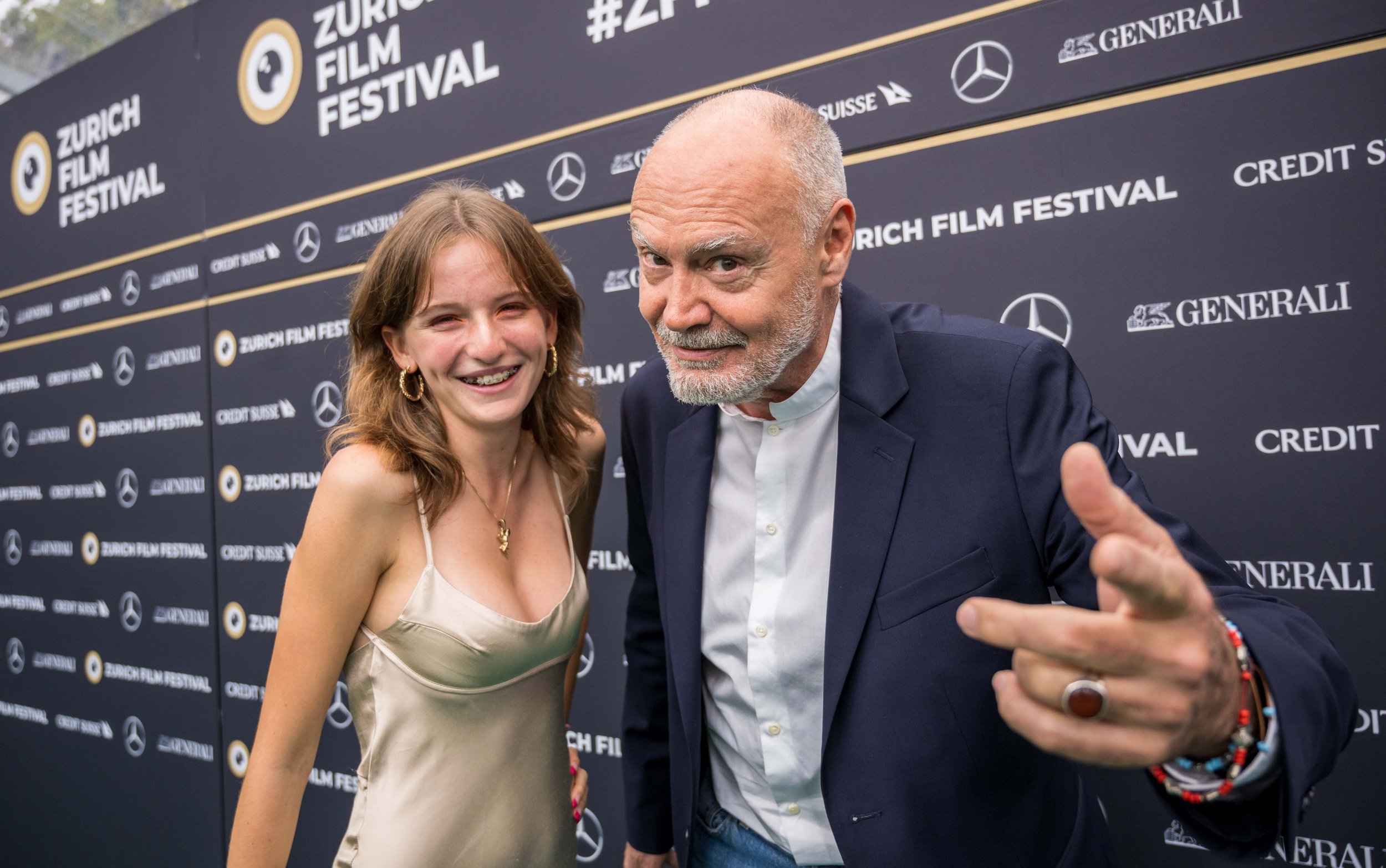 Two smiling people, a young woman and an older man, posing for a photo on the red carpet at the Zurich Film Festival, with a black backdrop featuring logos and the event name.