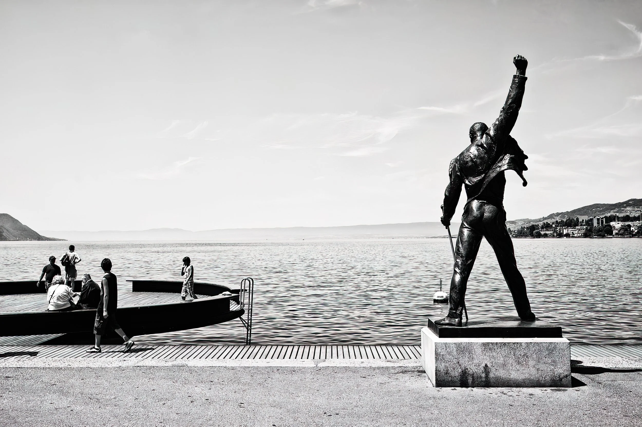 A crowd of people near a body of water with a large statue of a man holding a sword and raising his fist in the air, in front of a cityscape and hills.