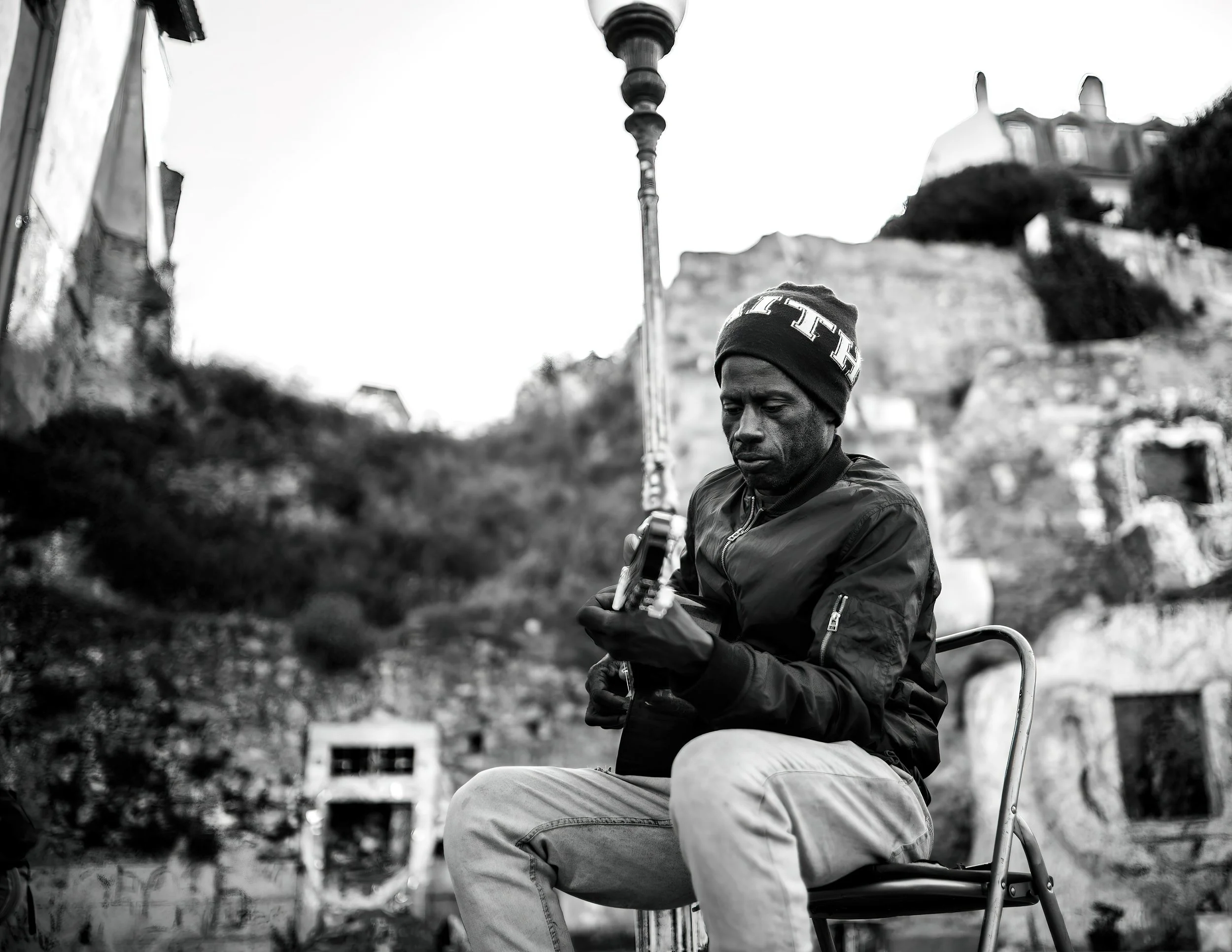 A man wearing a beanie and jacket sitting on a chair outdoors, playing a guitar.