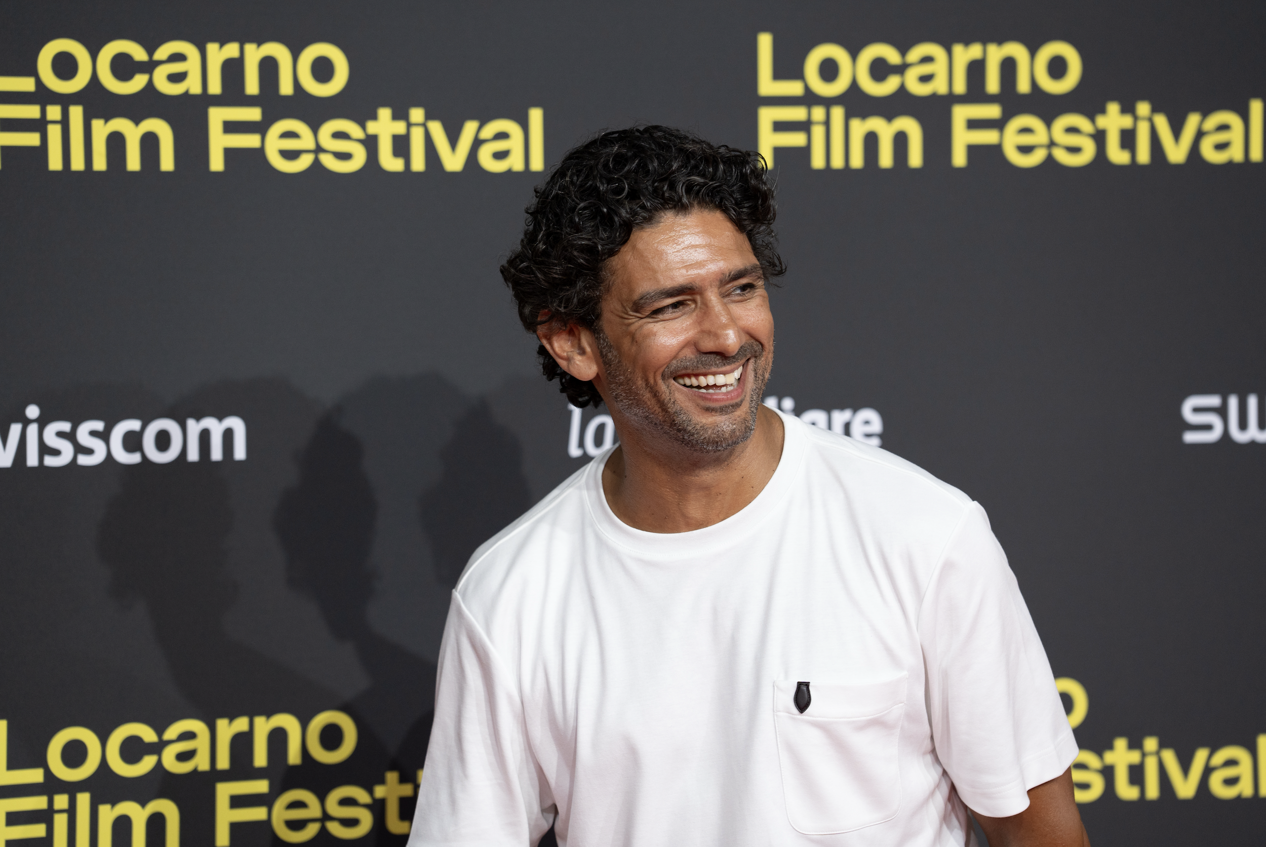 Man with dark curly hair and a beard smiling, wearing a white T-shirt, at the Locarno Film Festival.