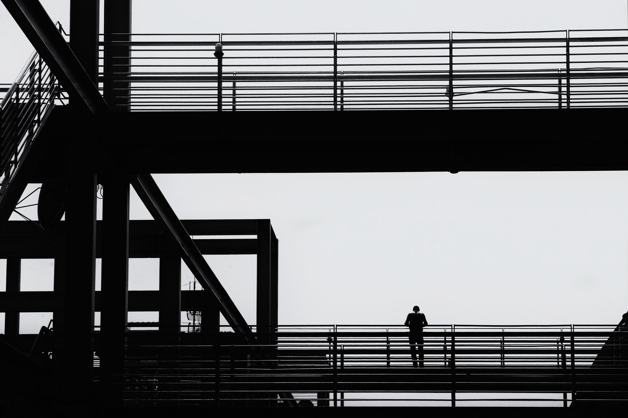 Silhouette of a person standing on a modern multi-level outdoor walkway with railings, with structural elements and more walkway levels in the background, against a cloudy sky.