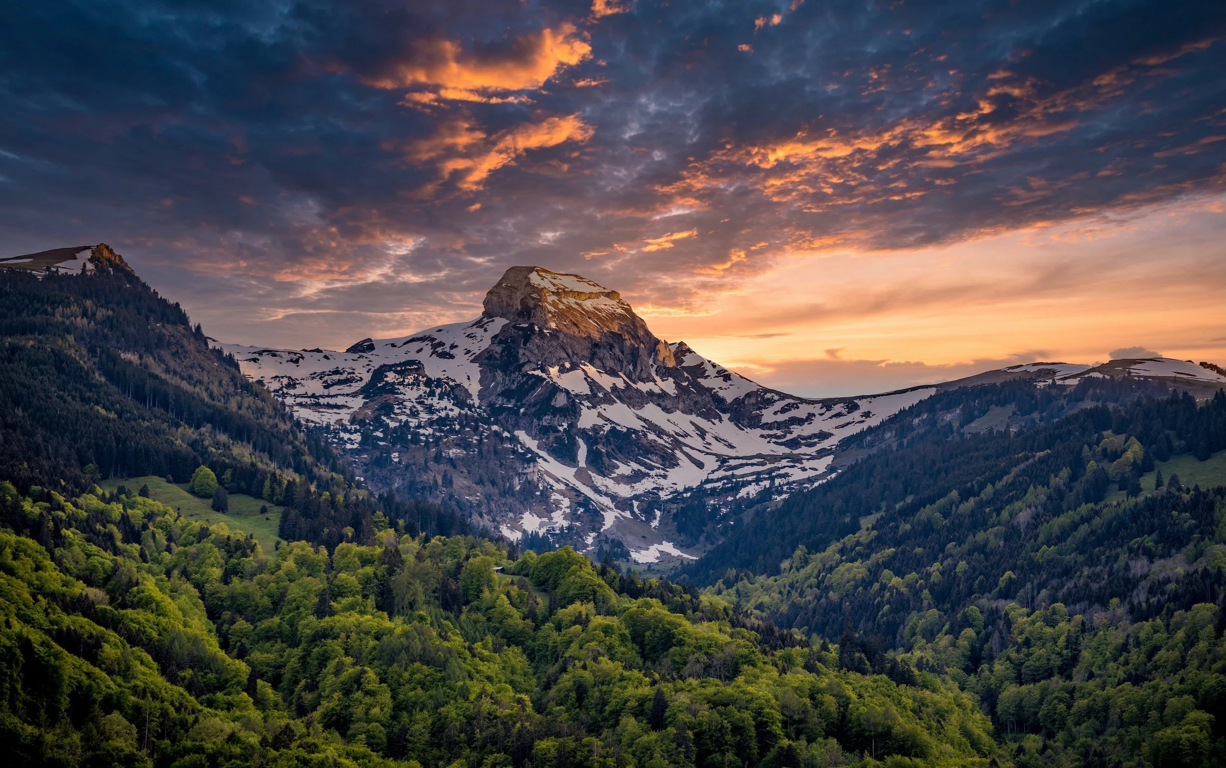 A mountain landscape view at sunset with snow-capped peaks and green forested hills.