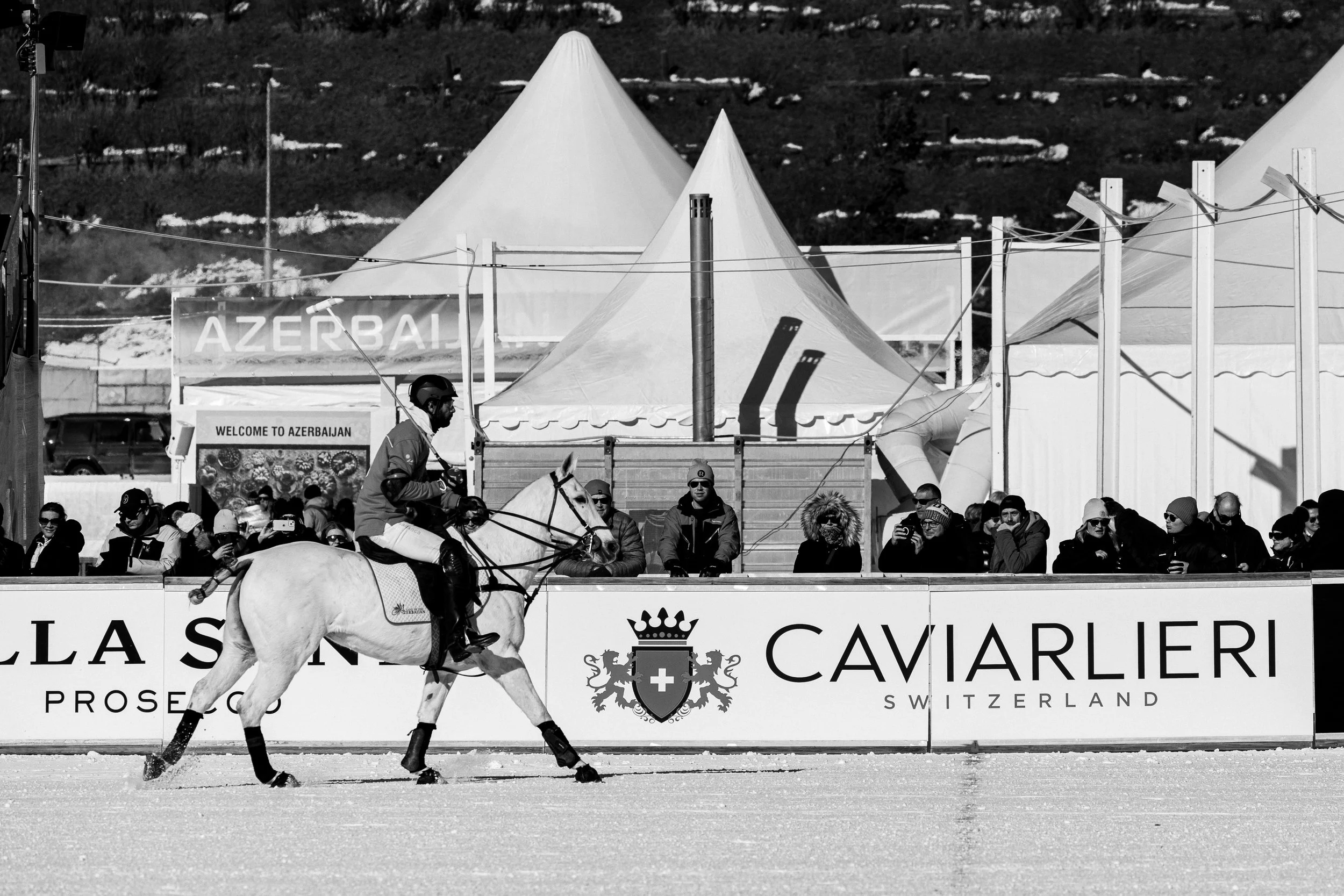 A man riding a white horse on a snow-covered surface at an event with a crowd of spectators behind a barrier. The backdrop features tents and a sign that reads "Welcome to Azerbaijan," with mountainous terrain in the distance.