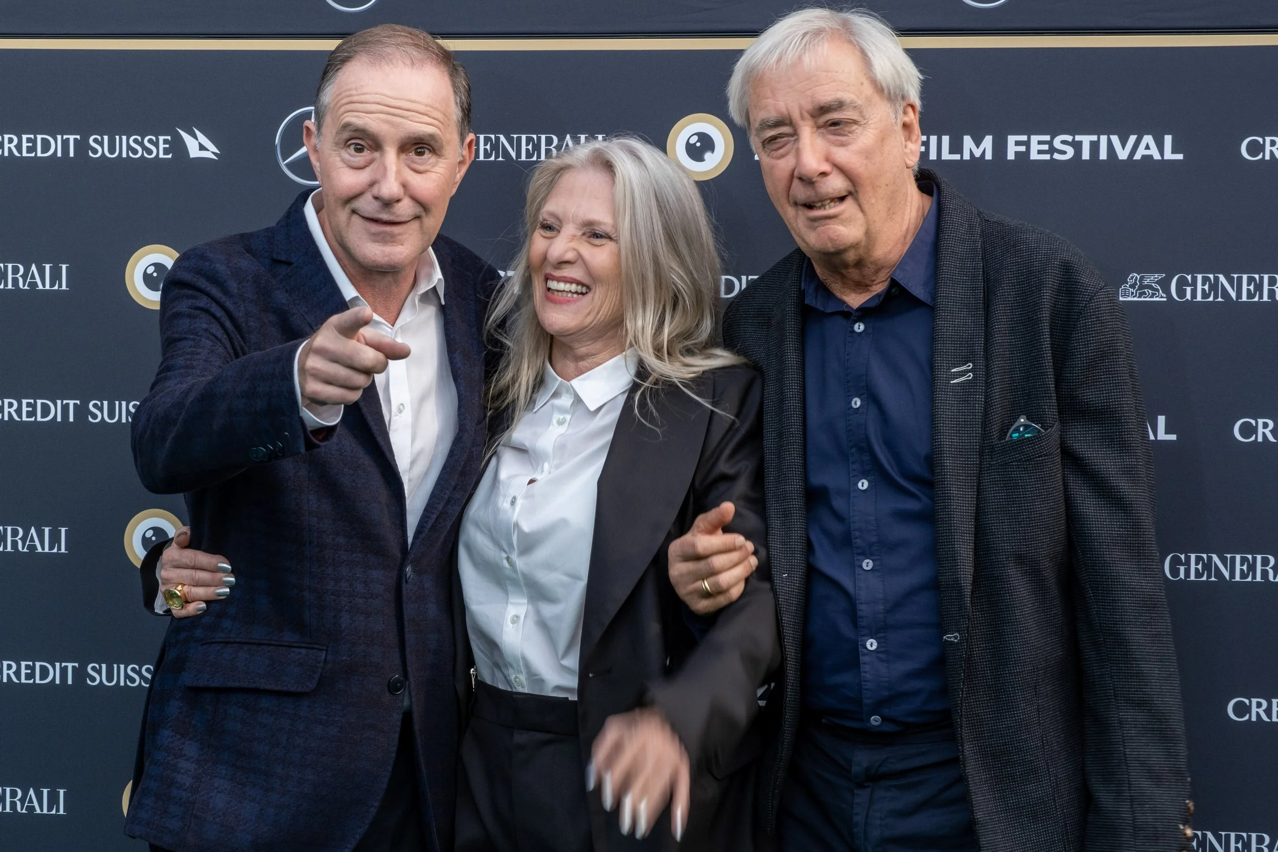 Three people standing together at a film festival, smiling and looking at the camera. The man on the left is pointing. The background has logos for Credit Suisse, Generali, and a film festival.
