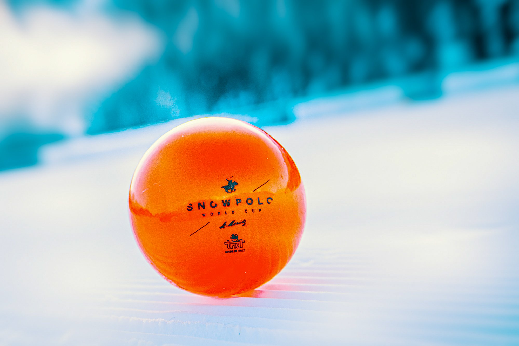An orange snowball with printed text and logos, placed on snow with a blurred winter landscape in the background.