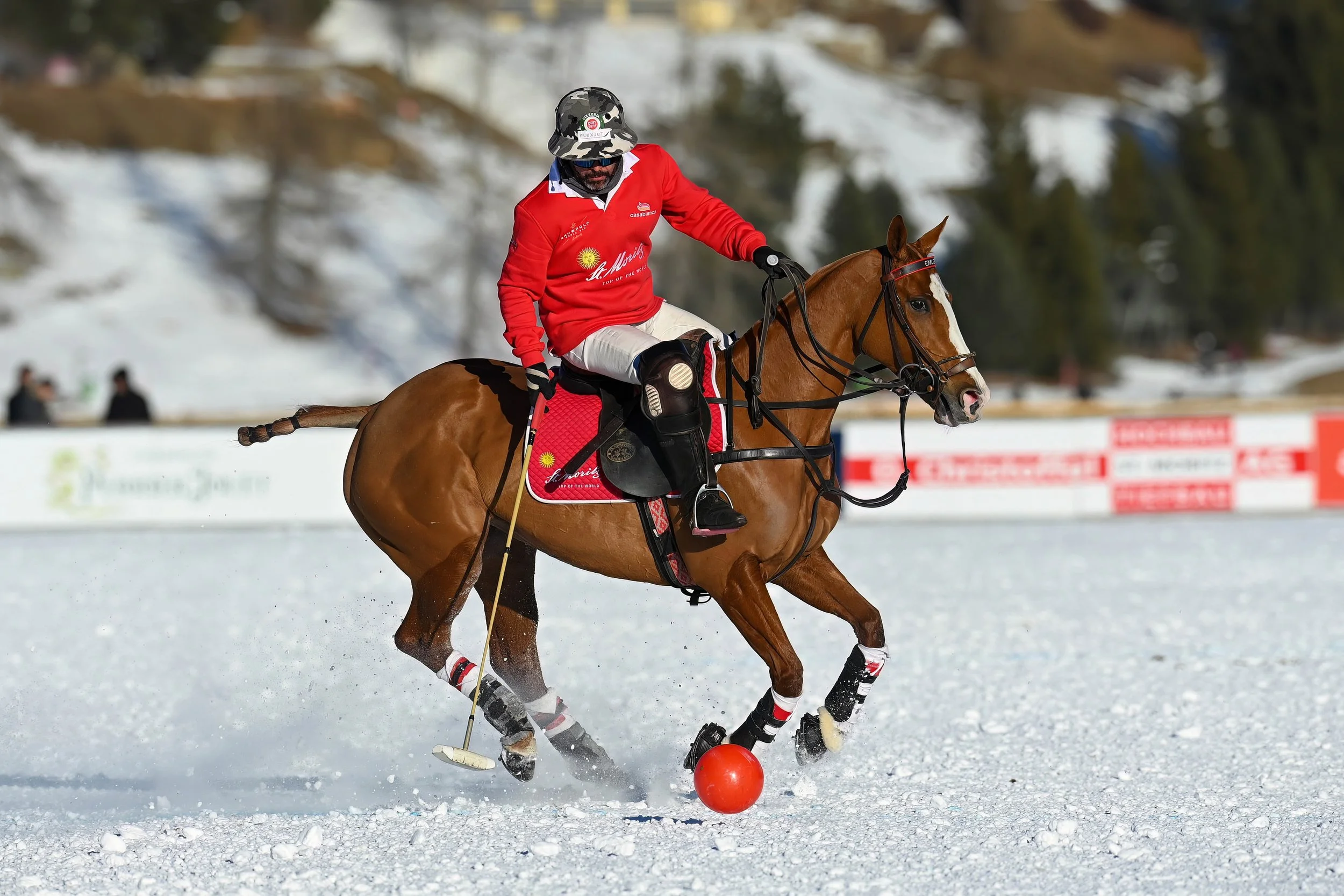 A polo player on horseback in a snowy field, hitting a red ball with a mallet during a game, wearing a red jersey and camouflage hat.