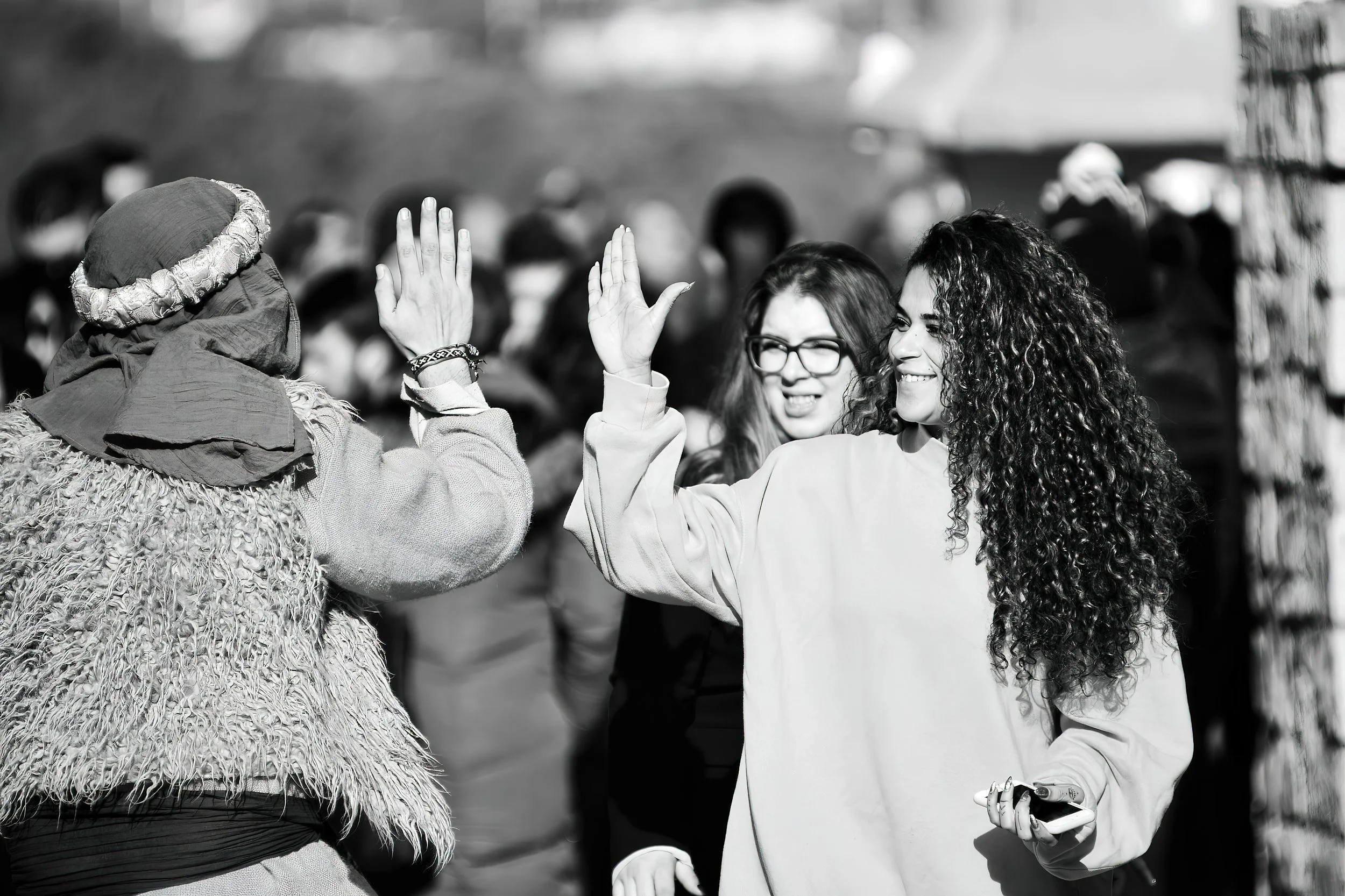 Two women with curly hair and glasses giving each other a high five in a crowded outdoor setting.