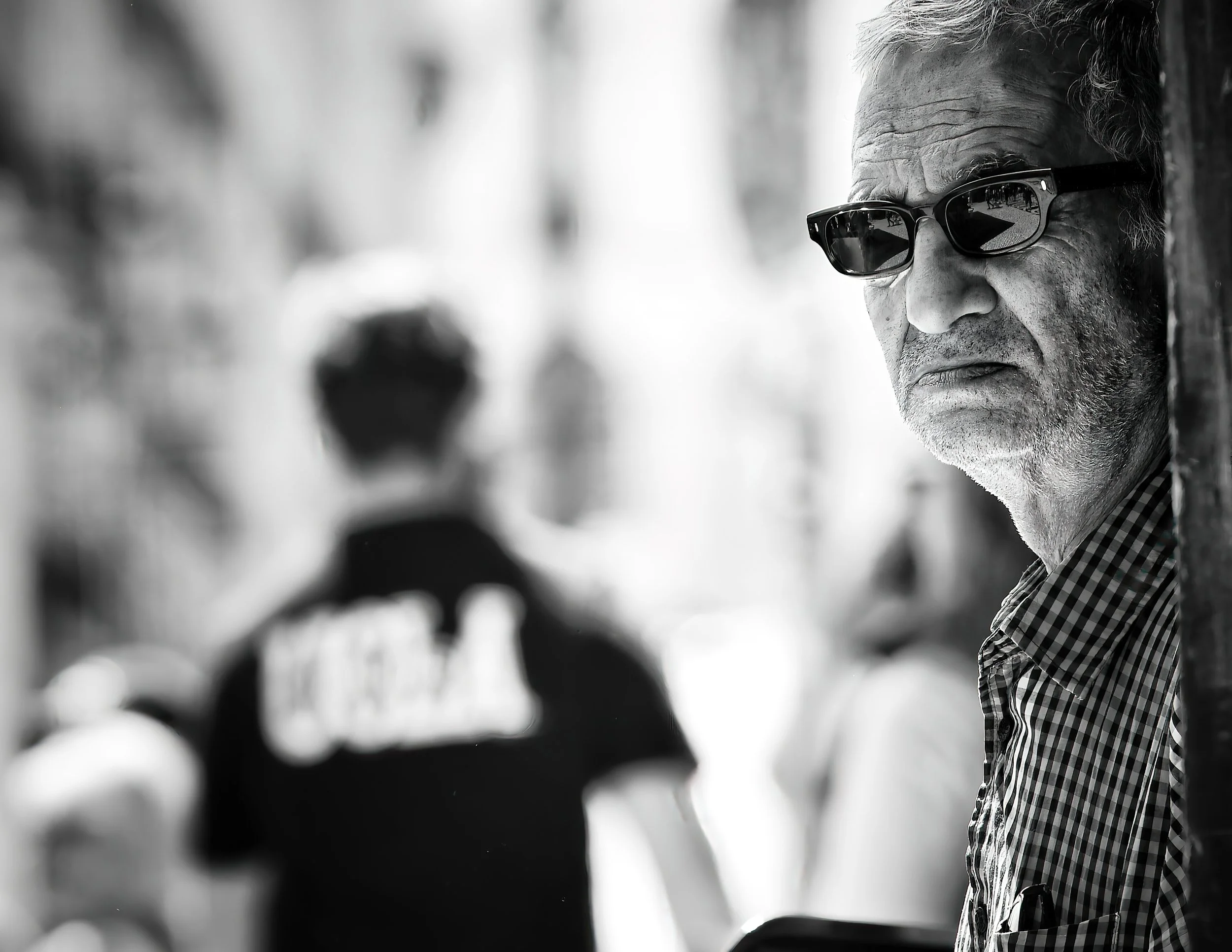 Older man wearing sunglasses and checkered shirt, looking serious, as he peers from behind a wall in a busy street scene, black and white photo.