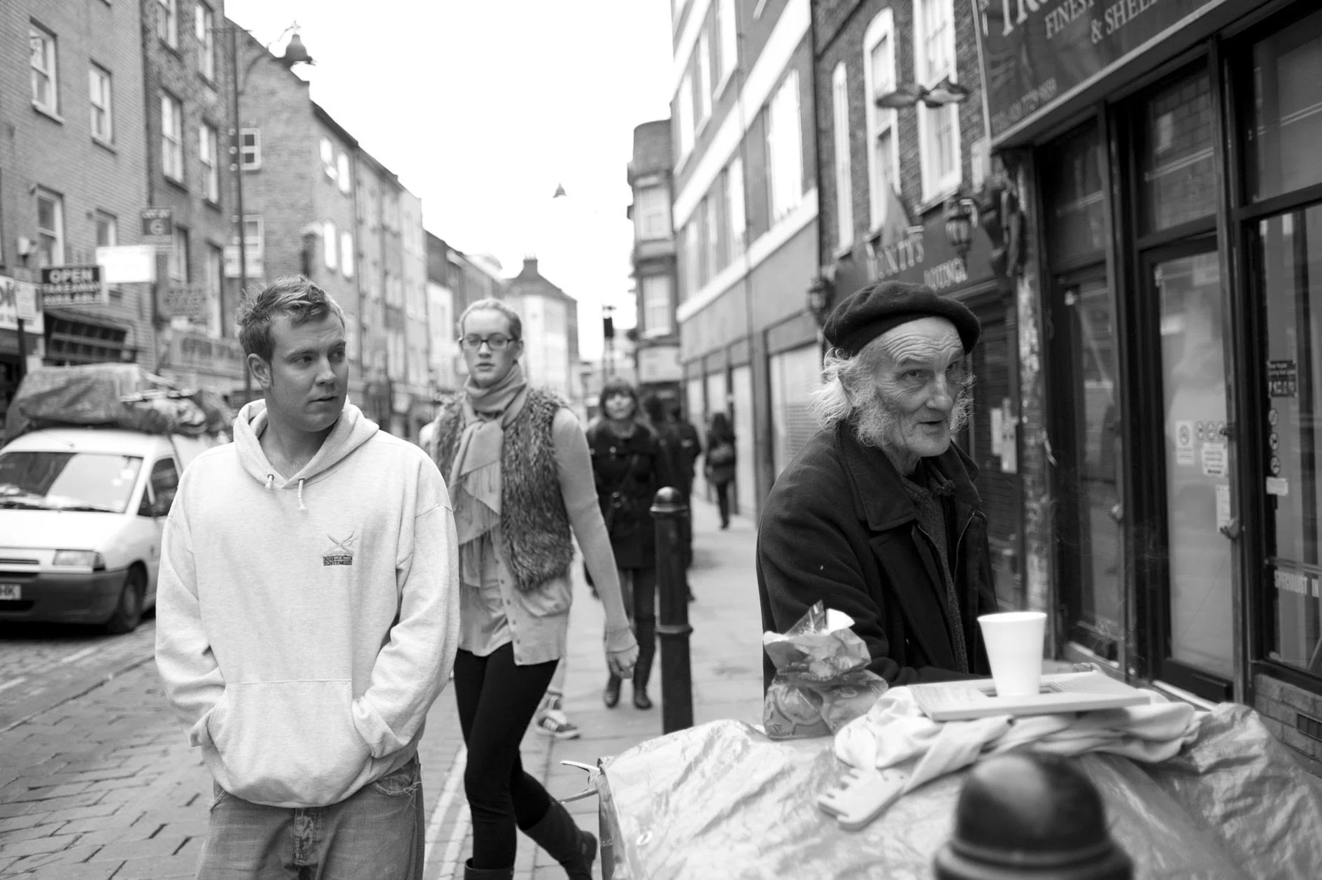 Street scene with three pedestrians walking past a street vendor selling food or drinks, in an urban area with brick and modern buildings, parked cars, and overhead streetlights.