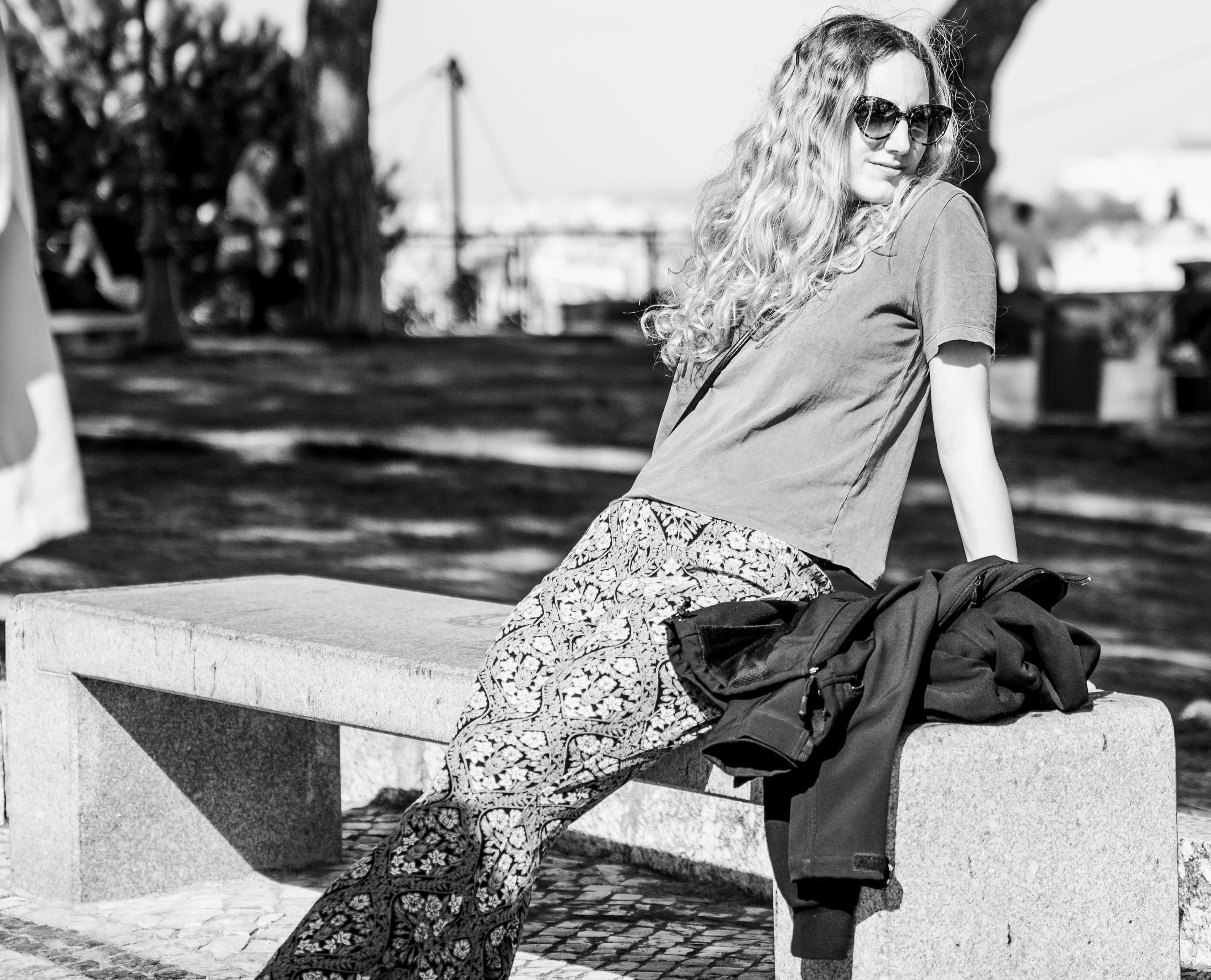 A woman with curly hair wearing sunglasses, a t-shirt, and patterned pants sits on a stone bench outdoors, with a jacket and bag beside her.