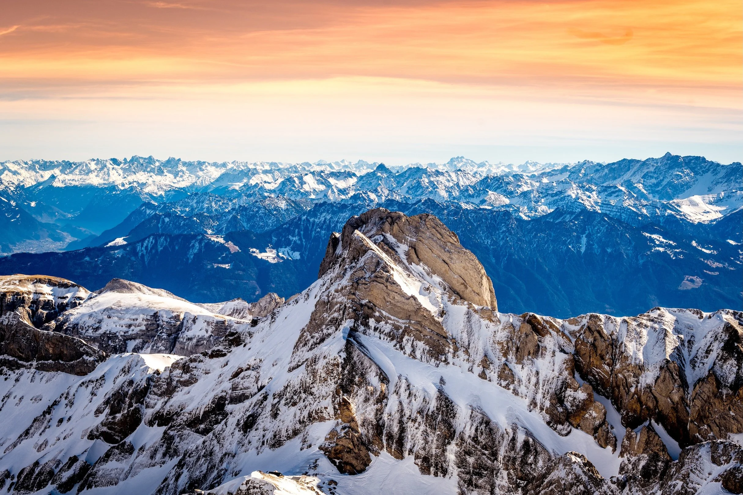 Snow-capped mountain peaks at sunset with layers of mountain ranges extending into the horizon.