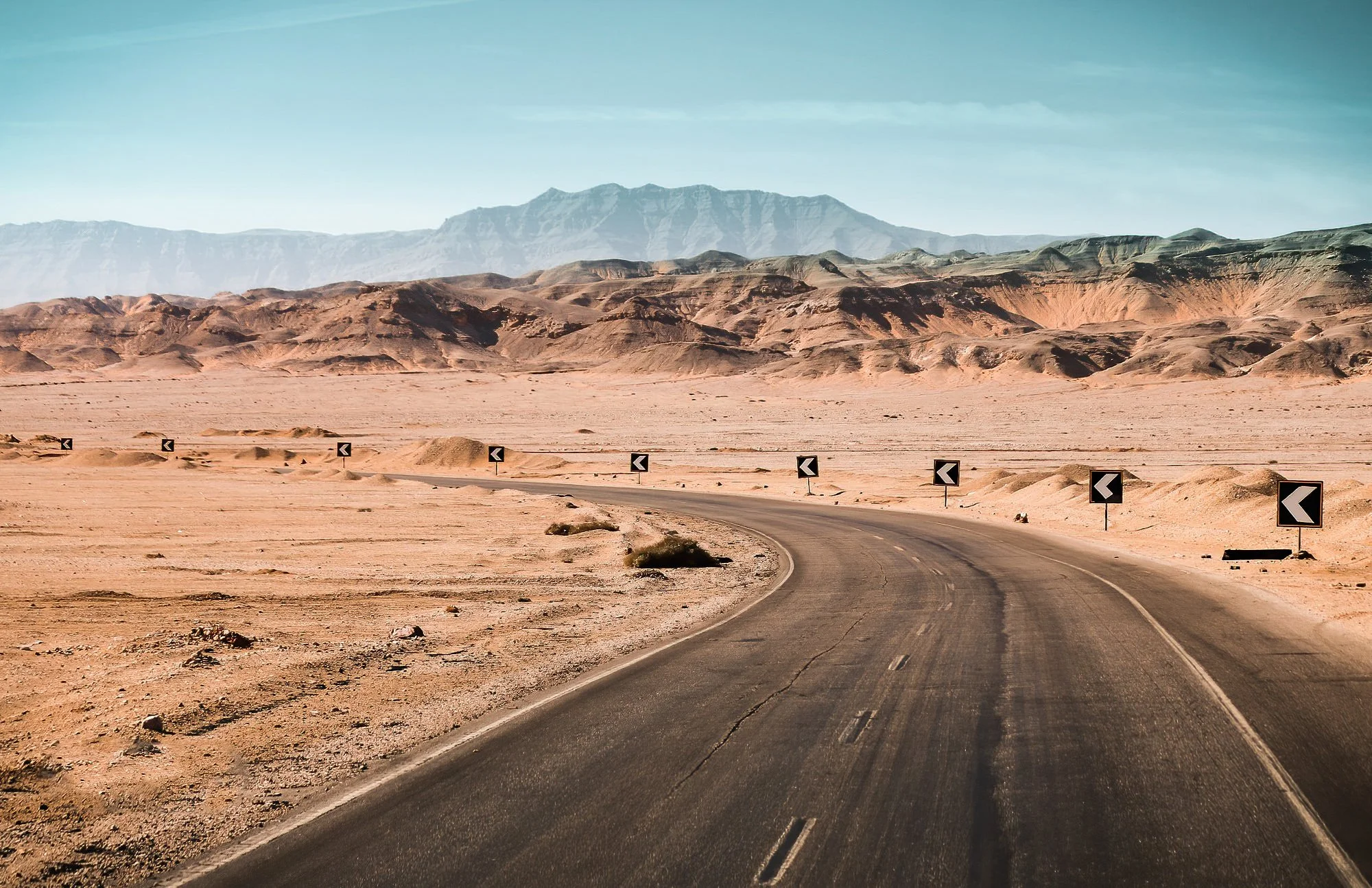 Winding road in a desert landscape with mountains in the background, directional chevron signs along the side of the road.
