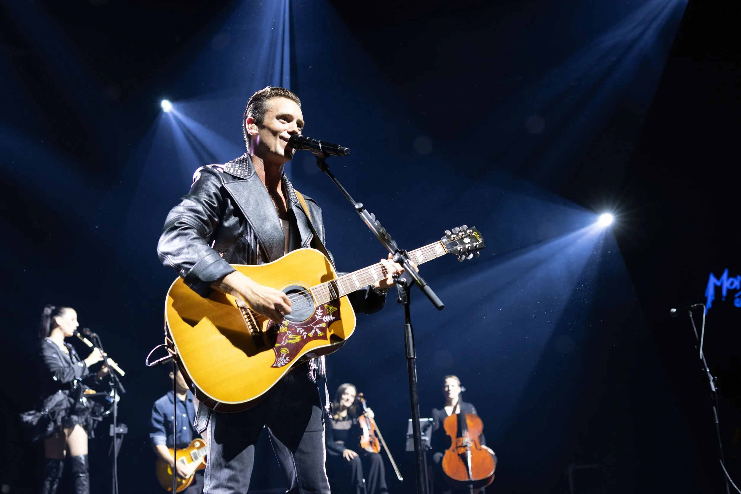 A man in a leather jacket playing an acoustic guitar and singing into a microphone on stage with spotlights shining down, accompanied by other musicians in the background.