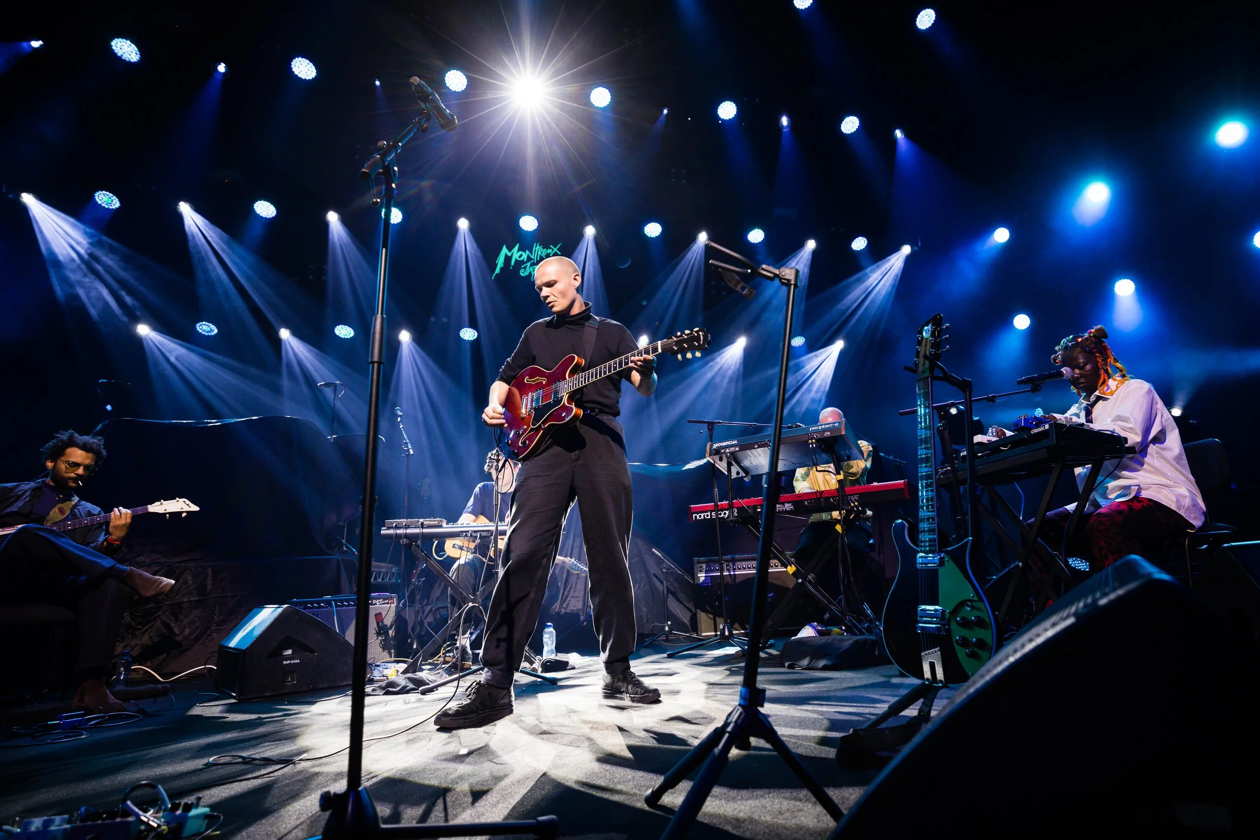 Musicians performing on stage under colorful lights, with a cue sign reading 'Montreux Jazz' in the background.