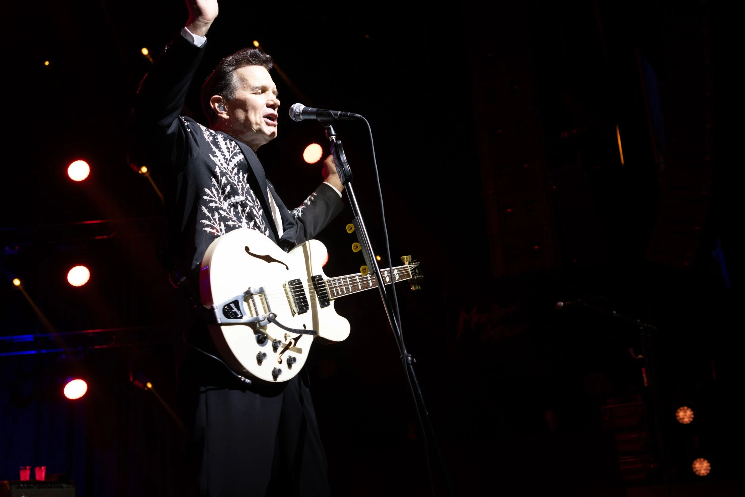 Performer singing passionately on stage with a white electric guitar, wearing a black suit with embroidered details, under stage lights.