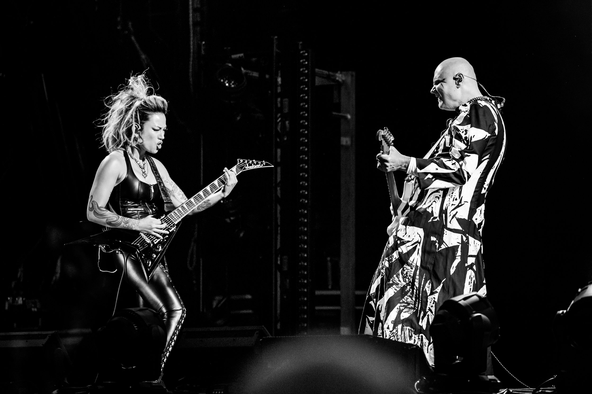 Two musicians perform on stage: a woman in leather pants and a sleeveless top playing an electric guitar, and a bald man in a patterned outfit playing a guitar, both passionately engaged in their performance.