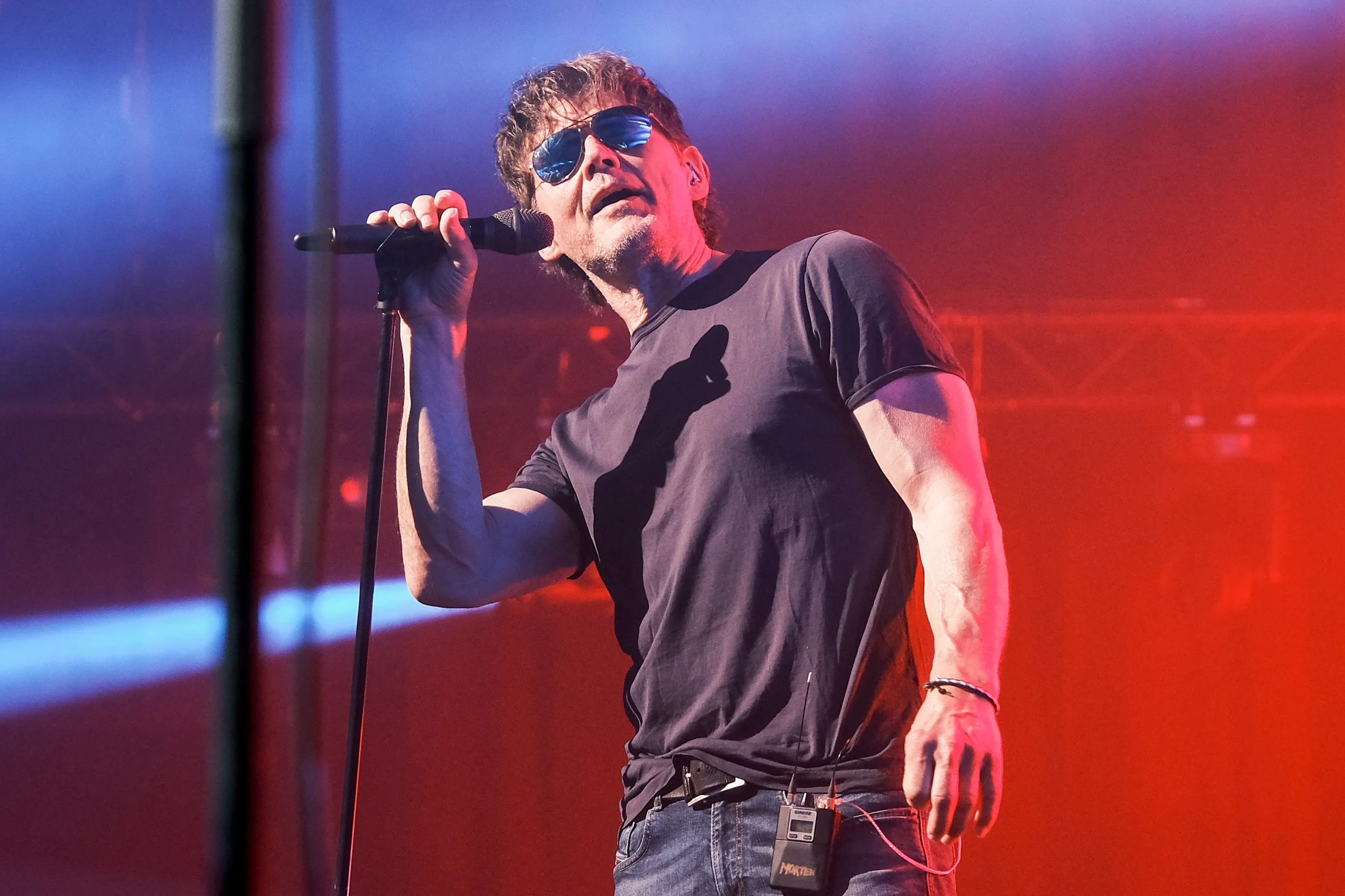Male singer performing on stage, wearing sunglasses, a black T-shirt, and holding a microphone.
