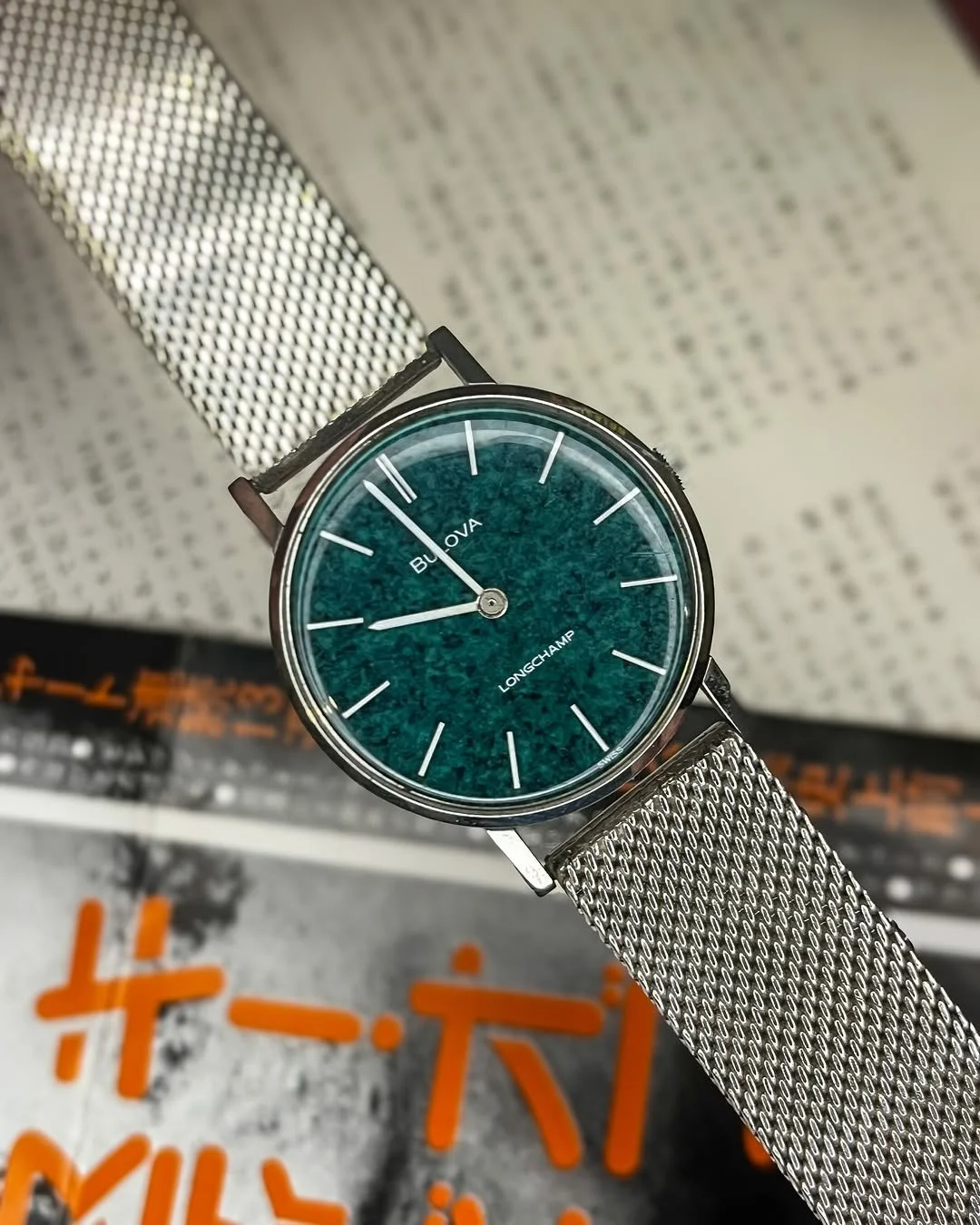A wristwatch with a teal jade stone dial, labeled 'Bulova' and 'Longchamp,' featuring minimalist white hour markers and a silver mesh band, resting on a blurred page with Japanese text.
