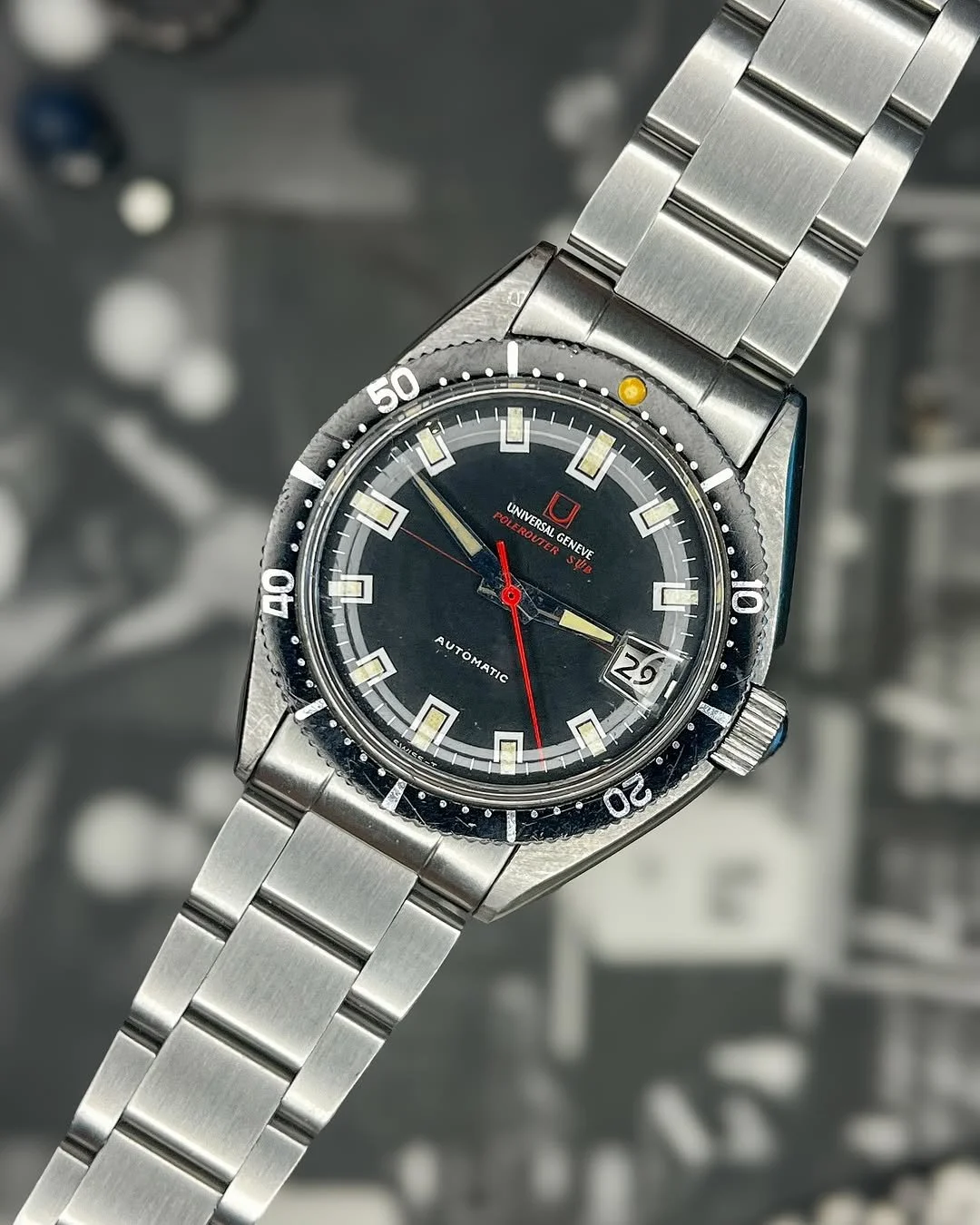 A vintage Universal Genève Polerouter Sub automatic wristwatch with a stainless steel bracelet, black dial, date window at 3 o'clock, rotating bezel, and chronograph features.