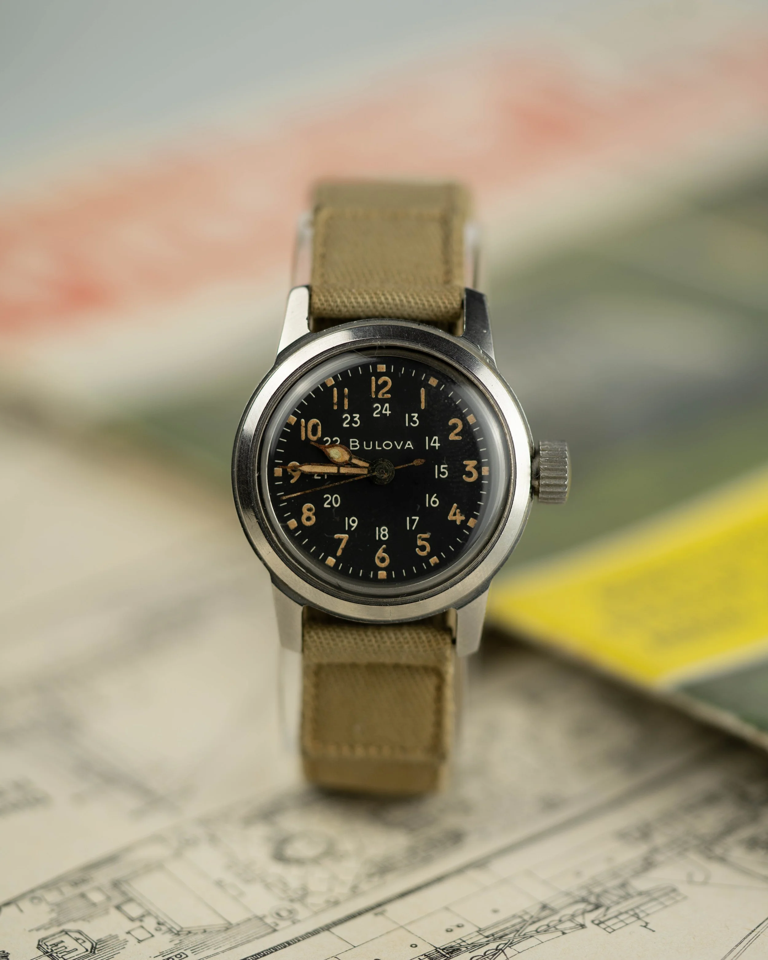 A vintage black dial US Military Issued Bulova wristwatch with a tan fabric strap, displayed on an architectural blueprint with blurred background.