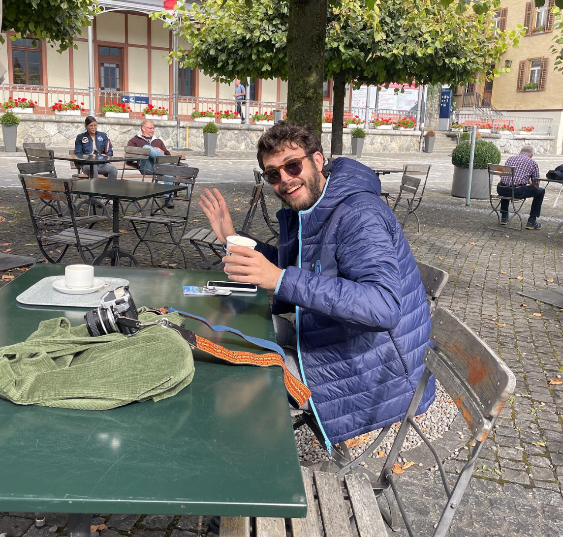 A man with sunglasses and a blue jacket sitting at an outdoor cafe table, smiling and holding a cup. On the table are a camera, a green bag, a smartphone, and a coffee cup.