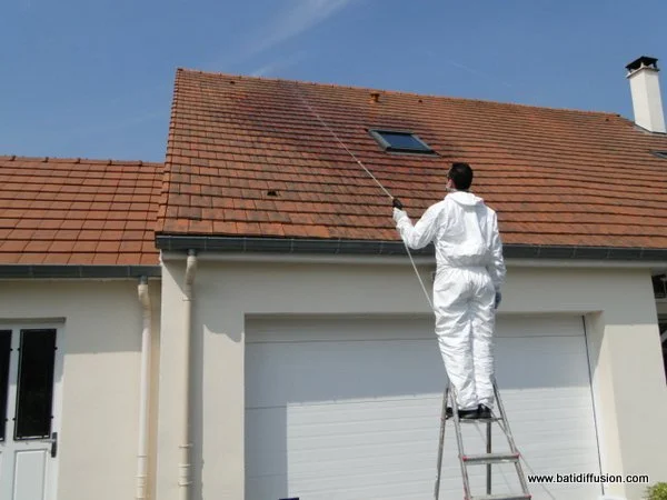 Un homme en combinaison blanche nettoie le toit d'une maison avec un nettoyeur à pression, utilisant une échelle.