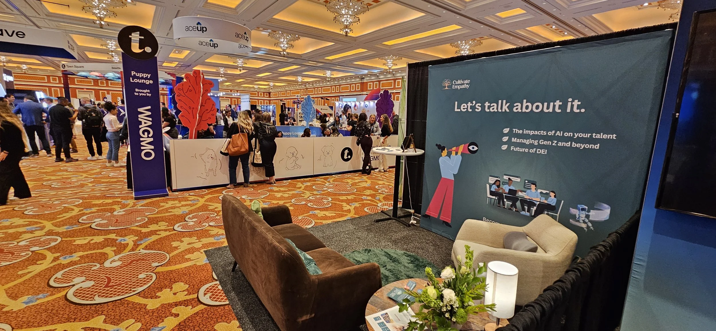 Trade show with a booth titled 'Cultivate Empathy' promoting discussions on AI, Gen Z, and future of DEI, featuring seating with flowers, large banners, and a busy background of attendees and booths.