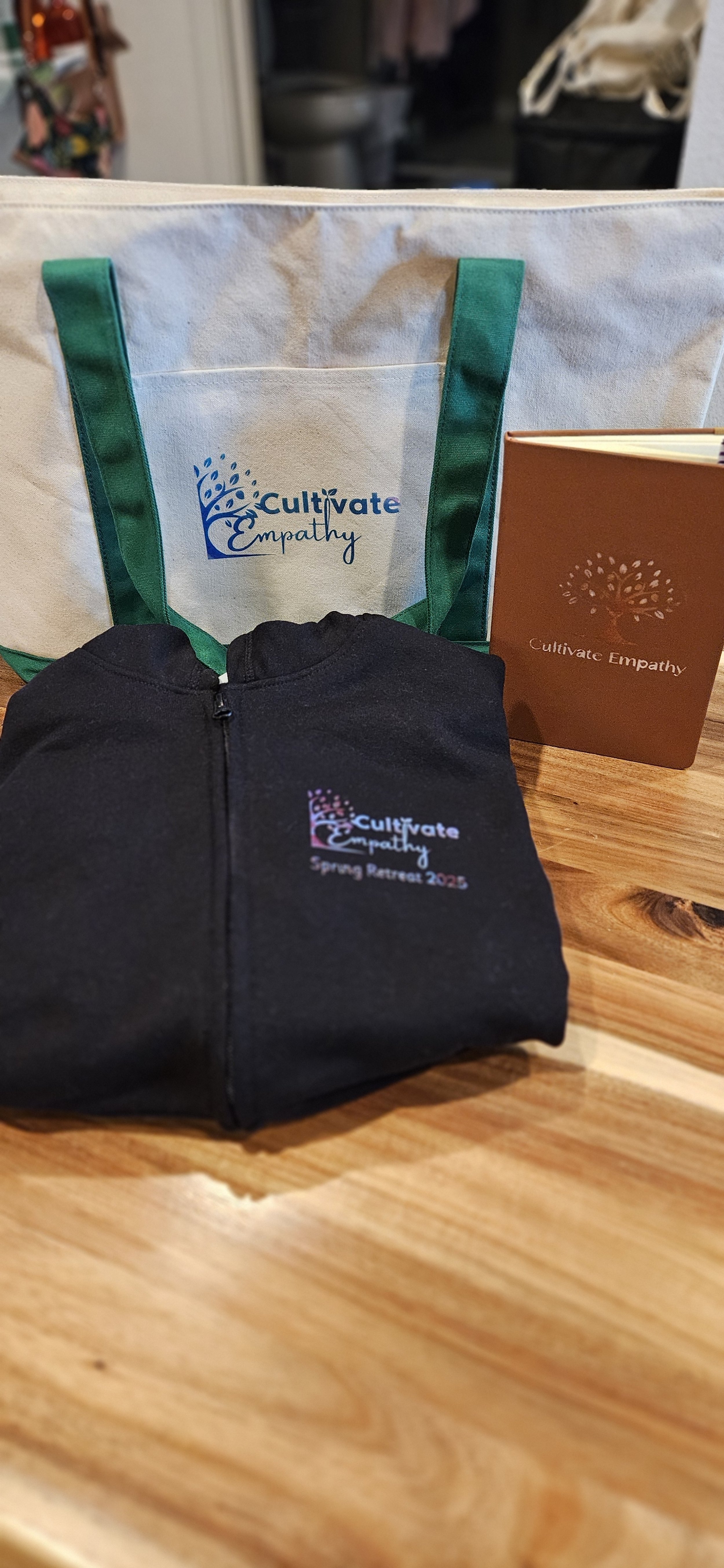 Folded black jacket with 'Cultivate Empathy Spring Retreat 2023' logo, display bag with logo, and brochure on a wooden table.