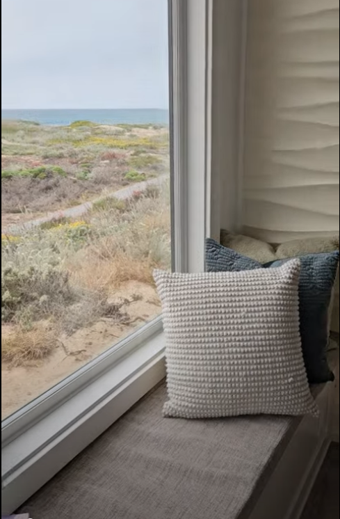 A pillow resting on a window seat next to a large window overlooking a beach with sand dunes, grass, and the ocean in the distance.