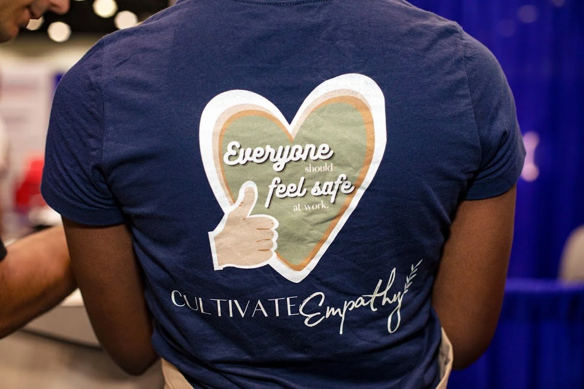 A person wearing a navy T-shirt that has a graphic of a heart with a thumbs-up inside it and the text "Everyone should feel safe at work" and "CULTIVATE Empathy" written on it.