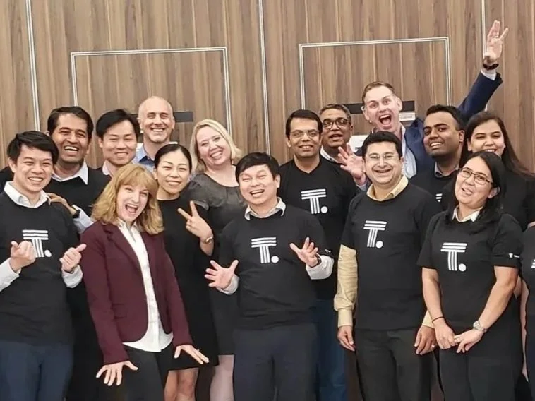 Group of diverse smiling people in business casual and casual attire posing together in a room with wooden walls, some wearing black shirts with a white 'T' and circular design.
