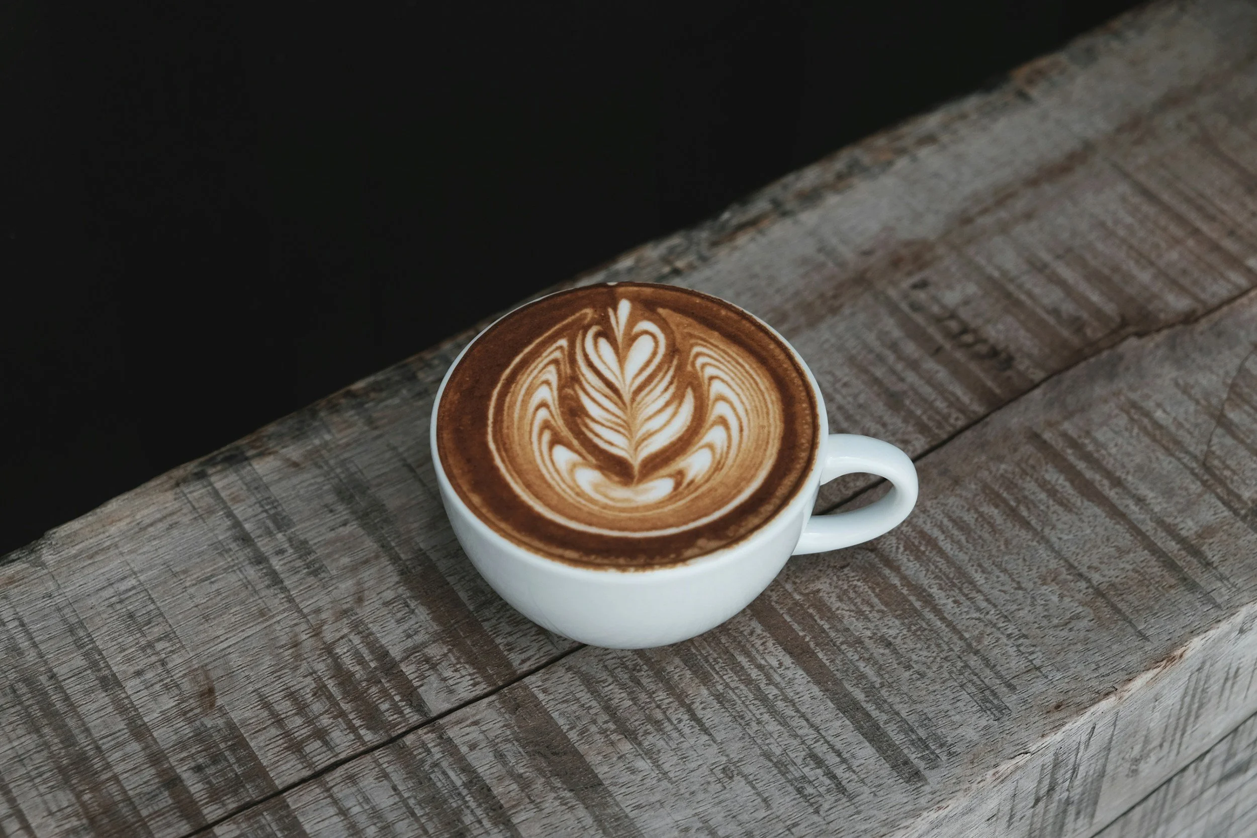 A white ceramic mug filled with a latte featuring latte art in the shape of a heart and leaves, placed on a rustic wooden table.