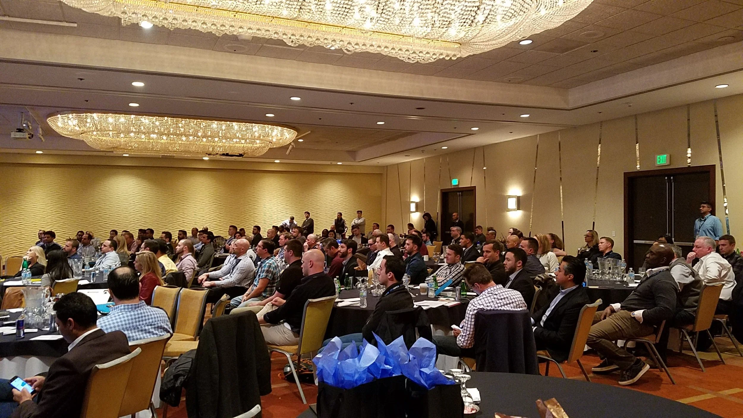 A large group of business professionals attending a conference in a hotel ballroom, sitting at tables with drinks and notebooks, attentively listening to a presentation.