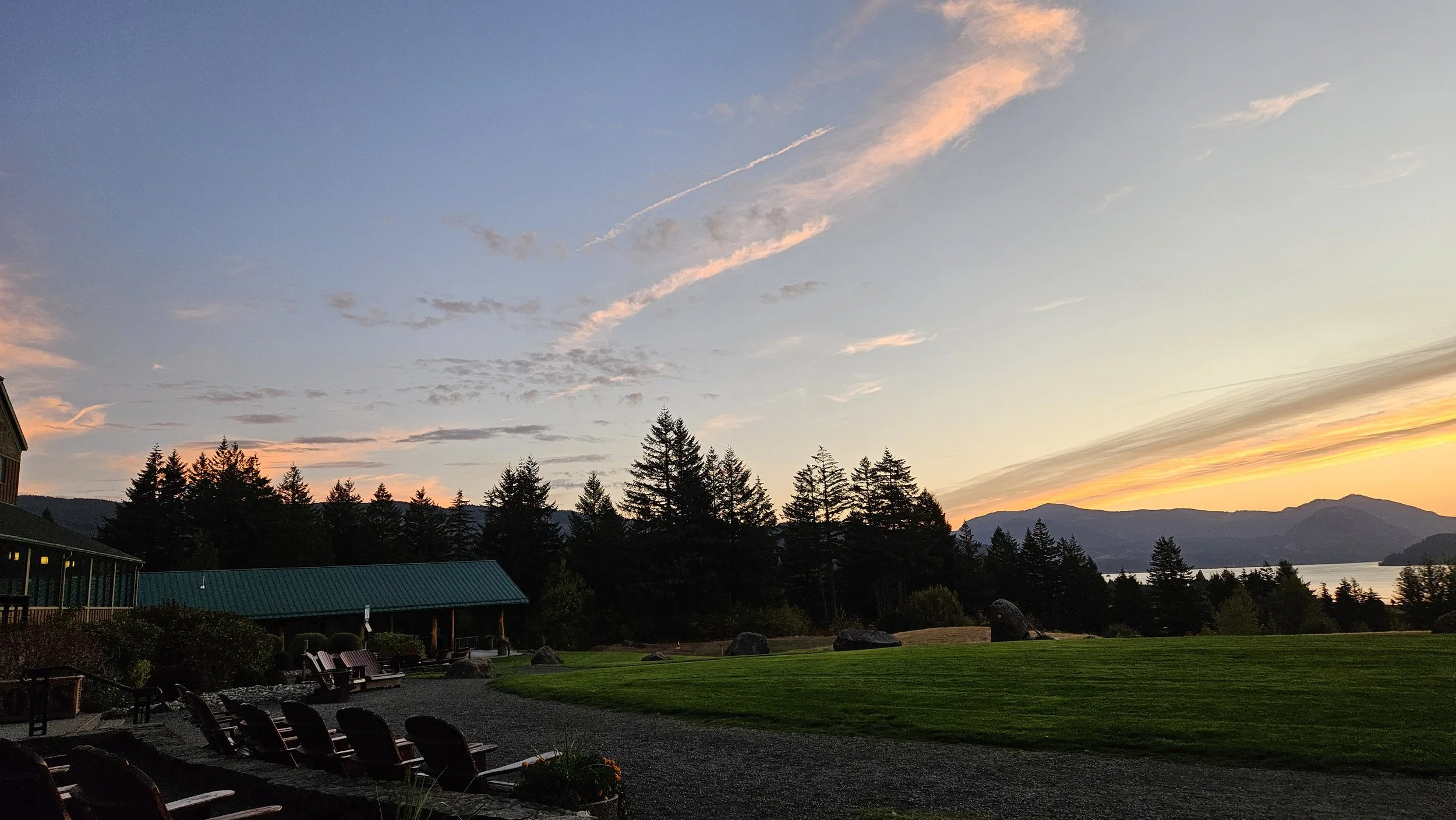 Sunset over lush green lawn with trees, mountains, and a lake in the distance; there are Adirondack chairs and a building with a green roof in the foreground.
