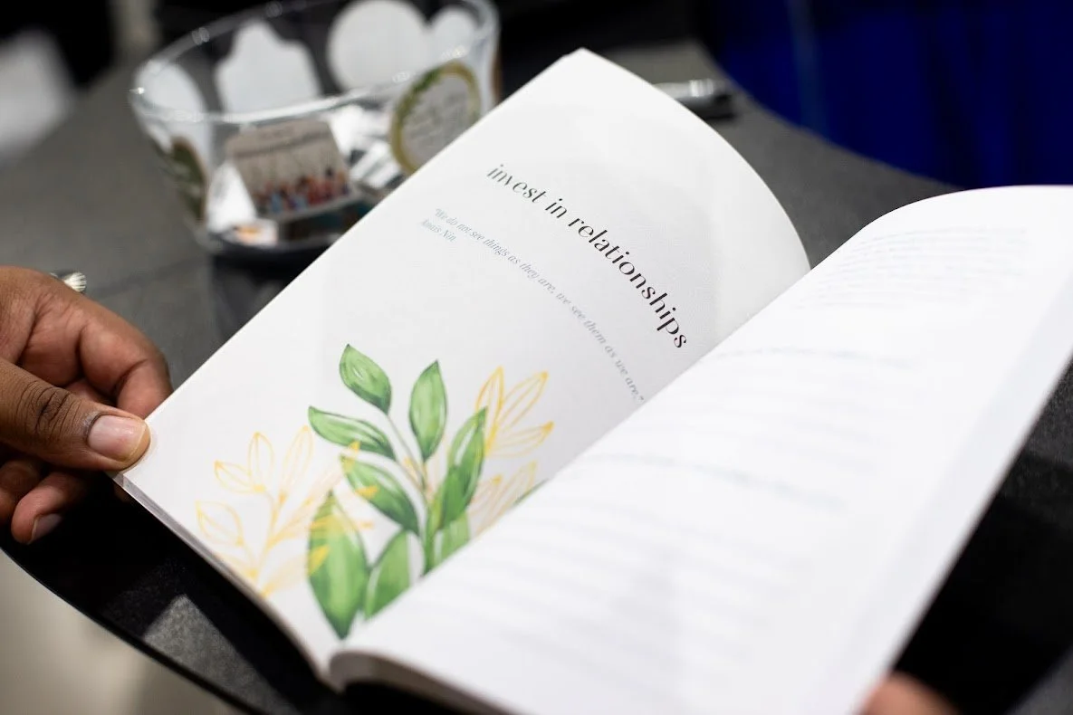 Open book with text about interest in relationships and a green leaf illustration, held by a person with dark skin, on a gray surface with a decorative bowl in the background.