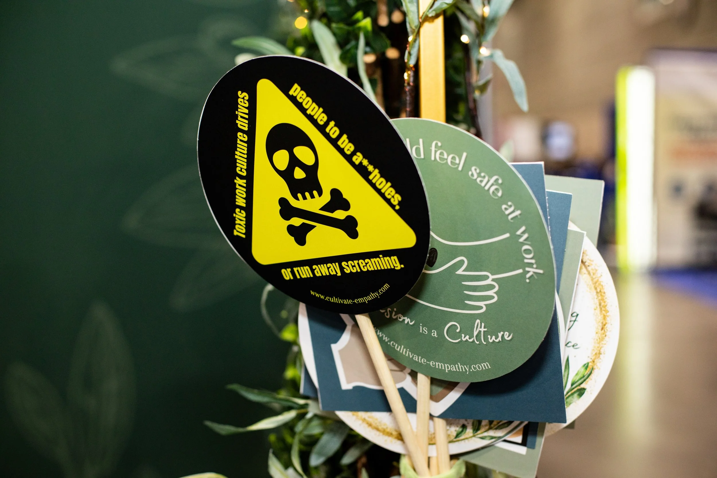 A collection of signs and flyers, one with a yellow and black warning design featuring a skull and crossbones and the text: "Toxic work culture drives people to be a**holes or run away screaming. www.cultivate-empathy.com."
