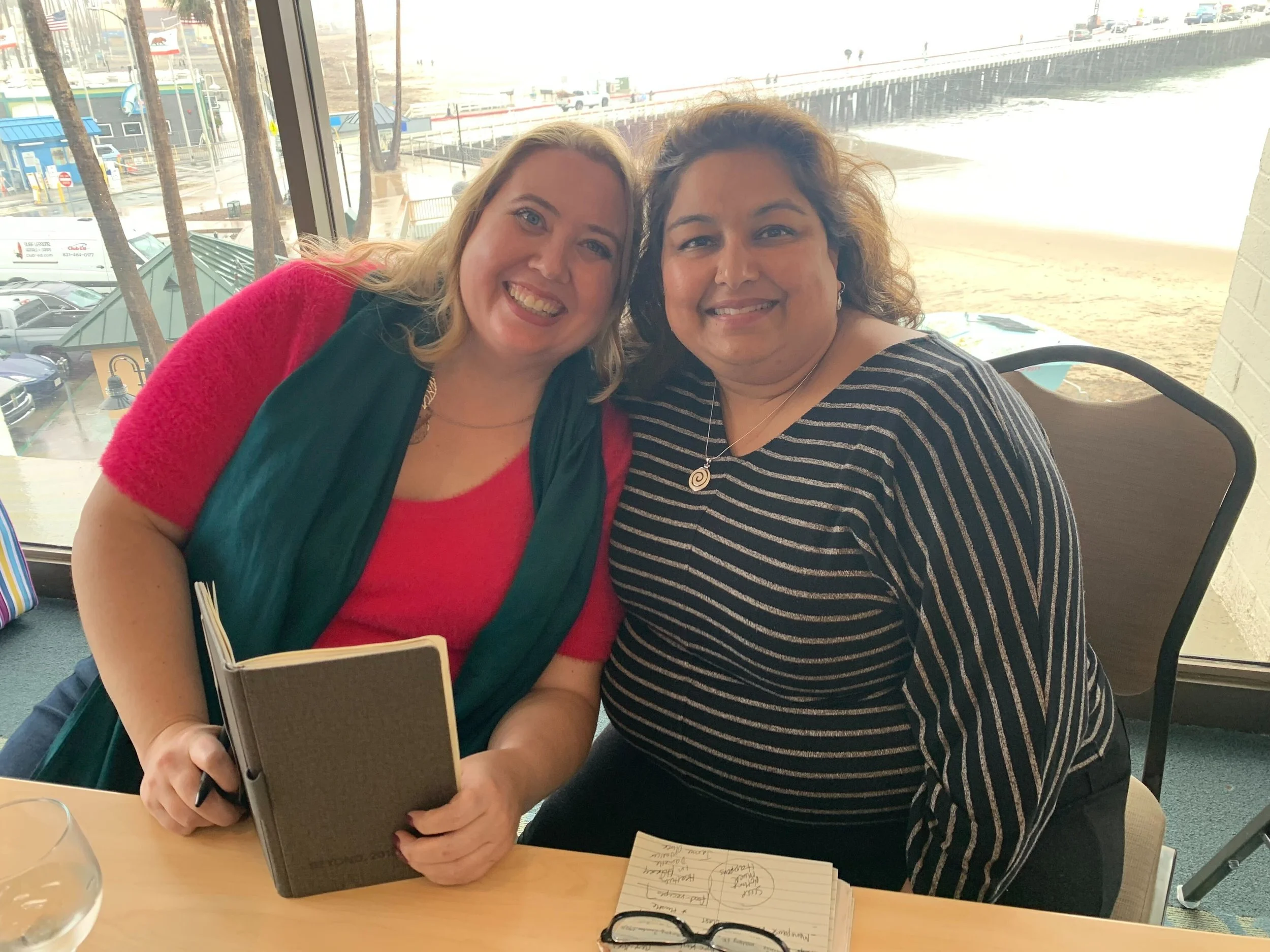 Two women smiling and sitting at a table near a window with an outdoor view of a beach and parking area.