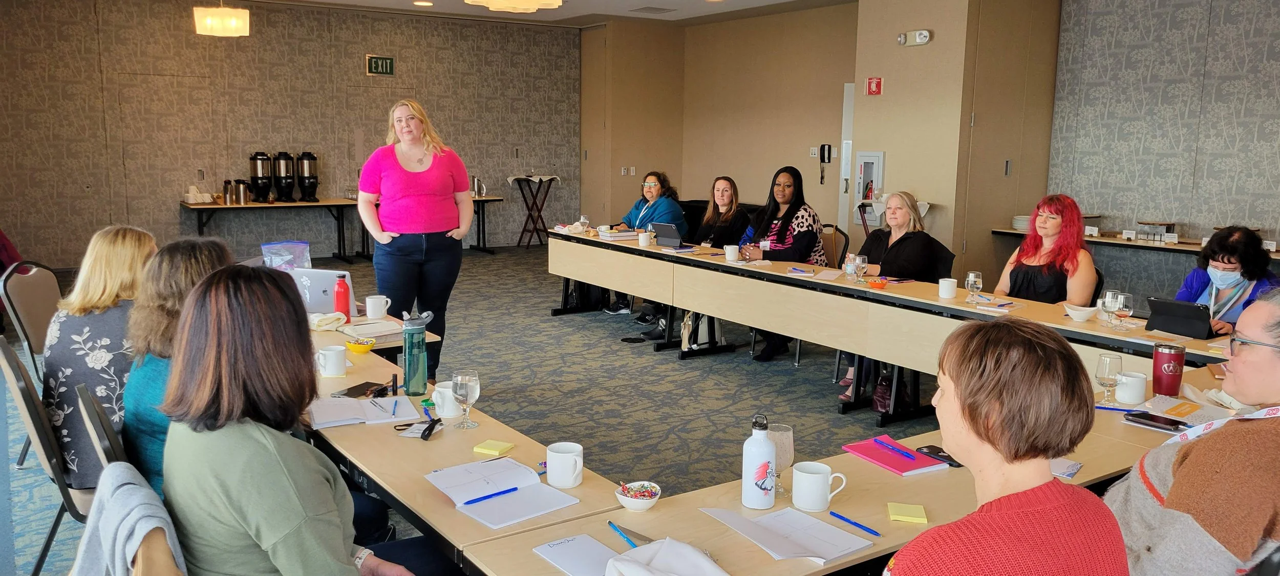 A woman in a pink top standing and speaking to a group of women seated around conference tables in a meeting room.