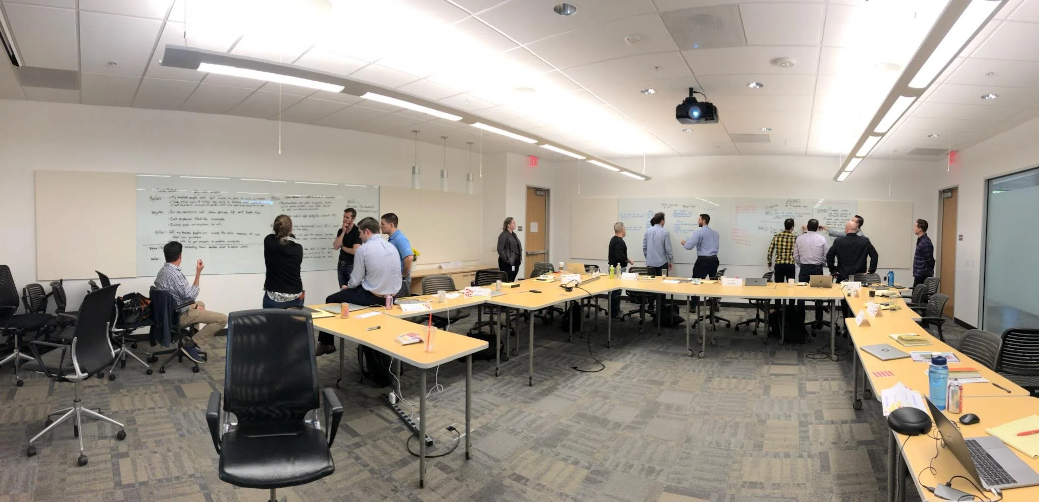 A conference room with multiple people standing and sitting around tables, whiteboards with notes on the walls, and a ceiling-mounted projector.