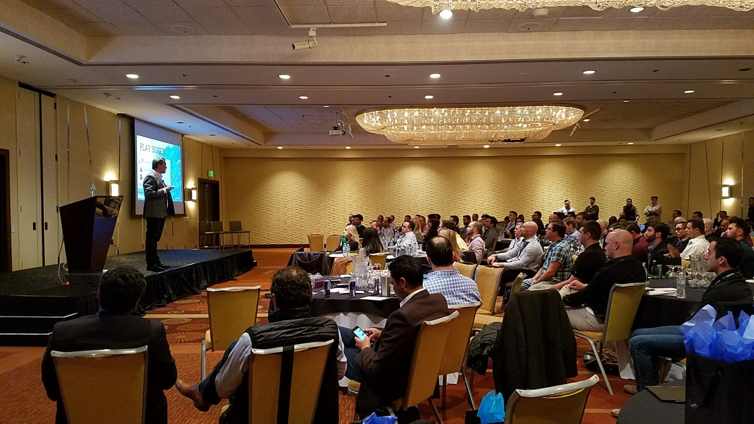 Business conference with a speaker on stage and audience seated at round tables in a hotel ballroom.
