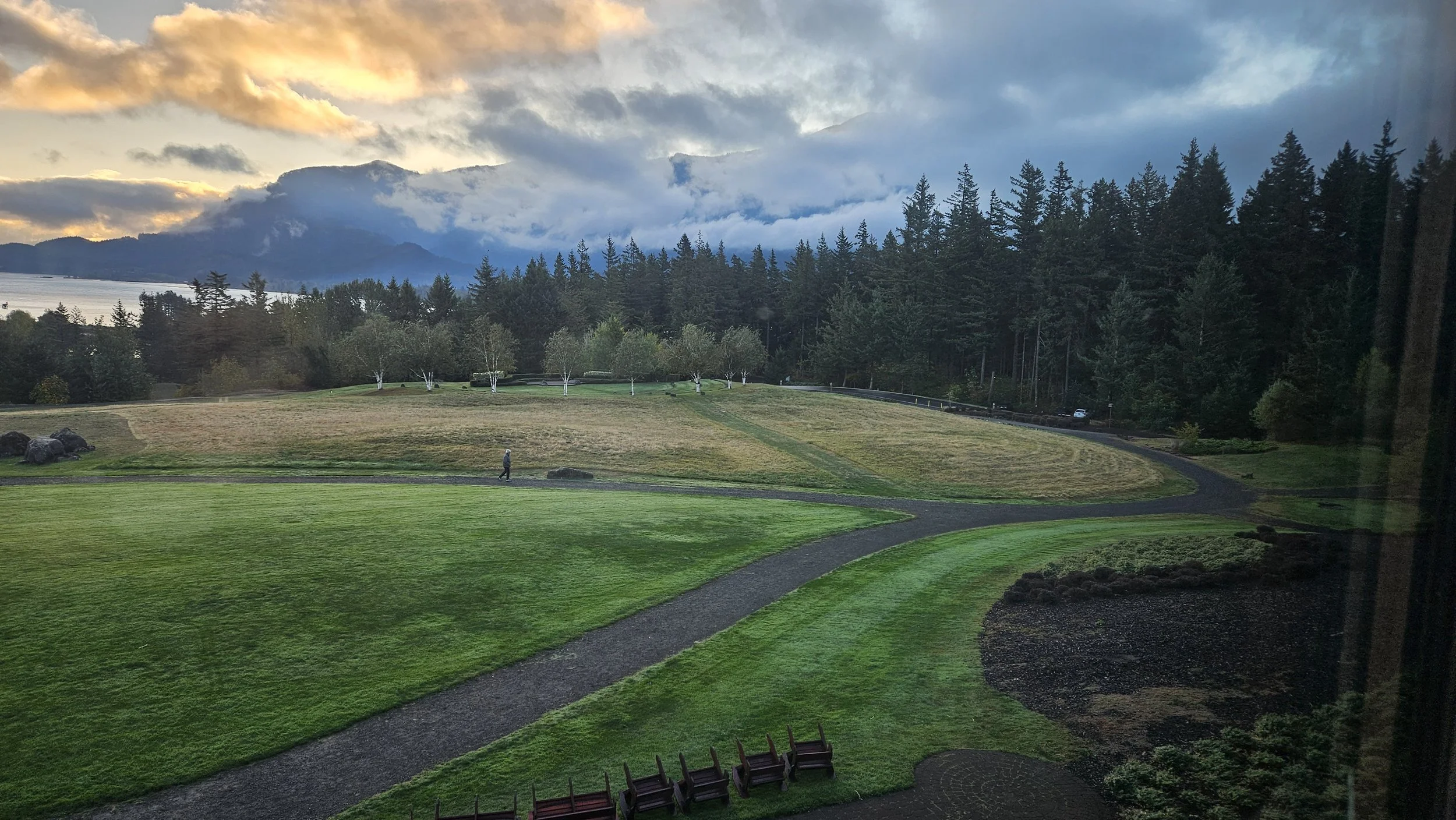 Scenic view of a park with a winding path, lush green grass, small trees, and a backdrop of mountains and cloudy sky at sunset.
