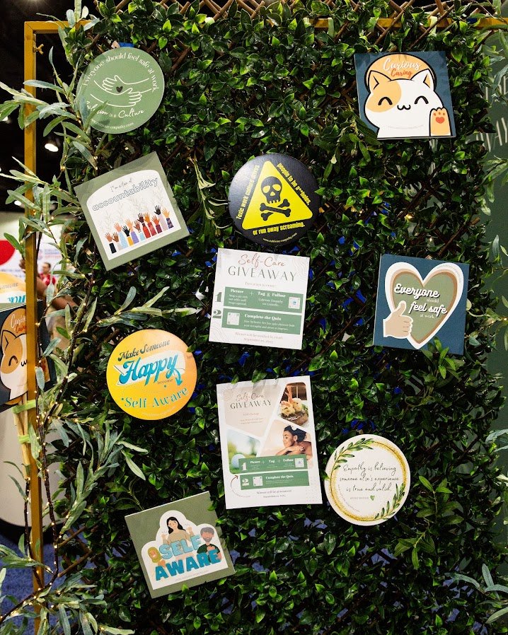 Green leafy wall decorated with various signs promoting self-care, mental health, and safety, including messages like "Everyone feels safe," "Make Someone Happy," and several giveaway notices.