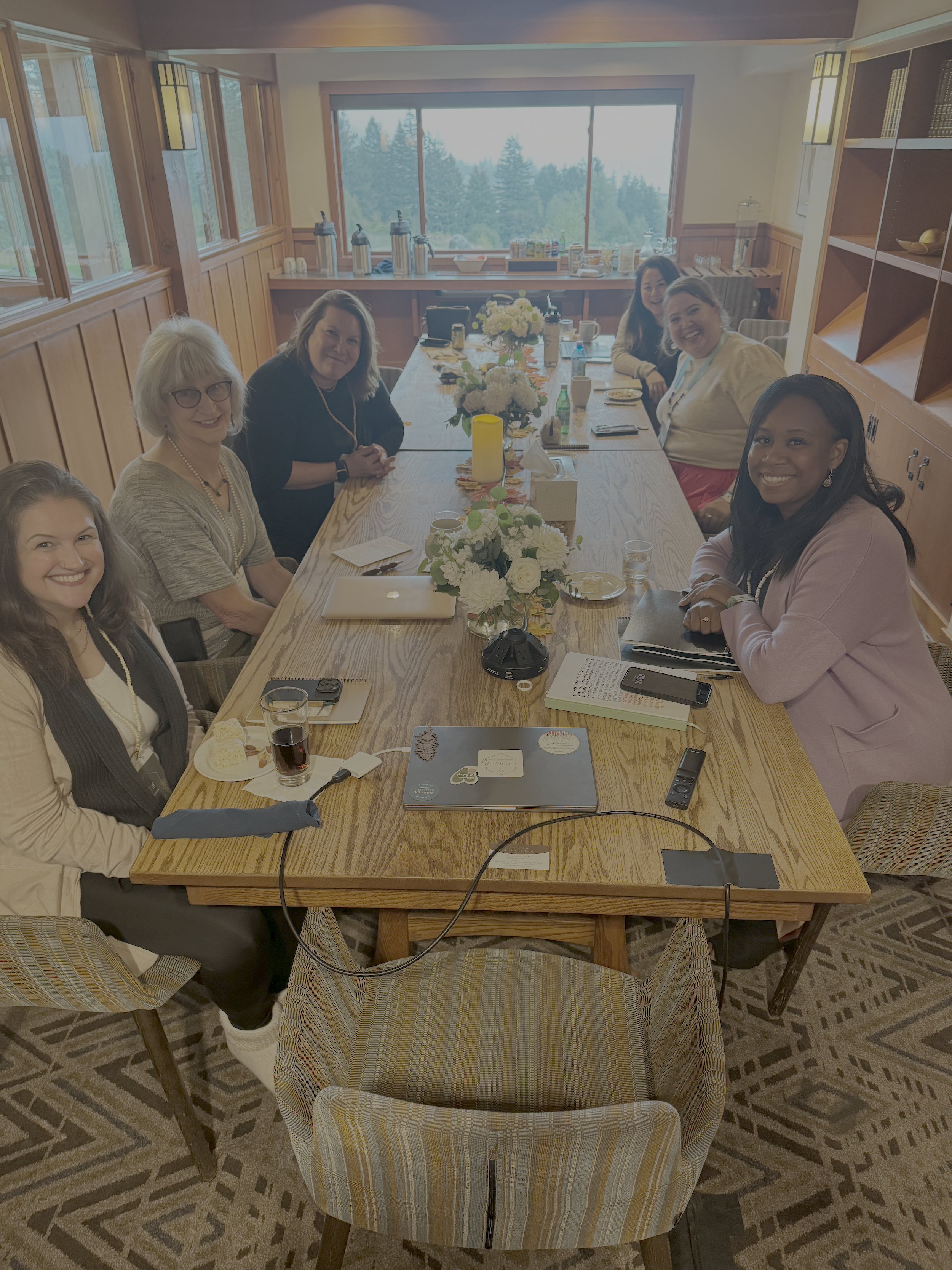 Seven women sitting around a wooden conference table in a well-lit room with large window and wooden paneling. The table has flowers, drinks, notebooks, and electronic devices, and the women are smiling at the camera.