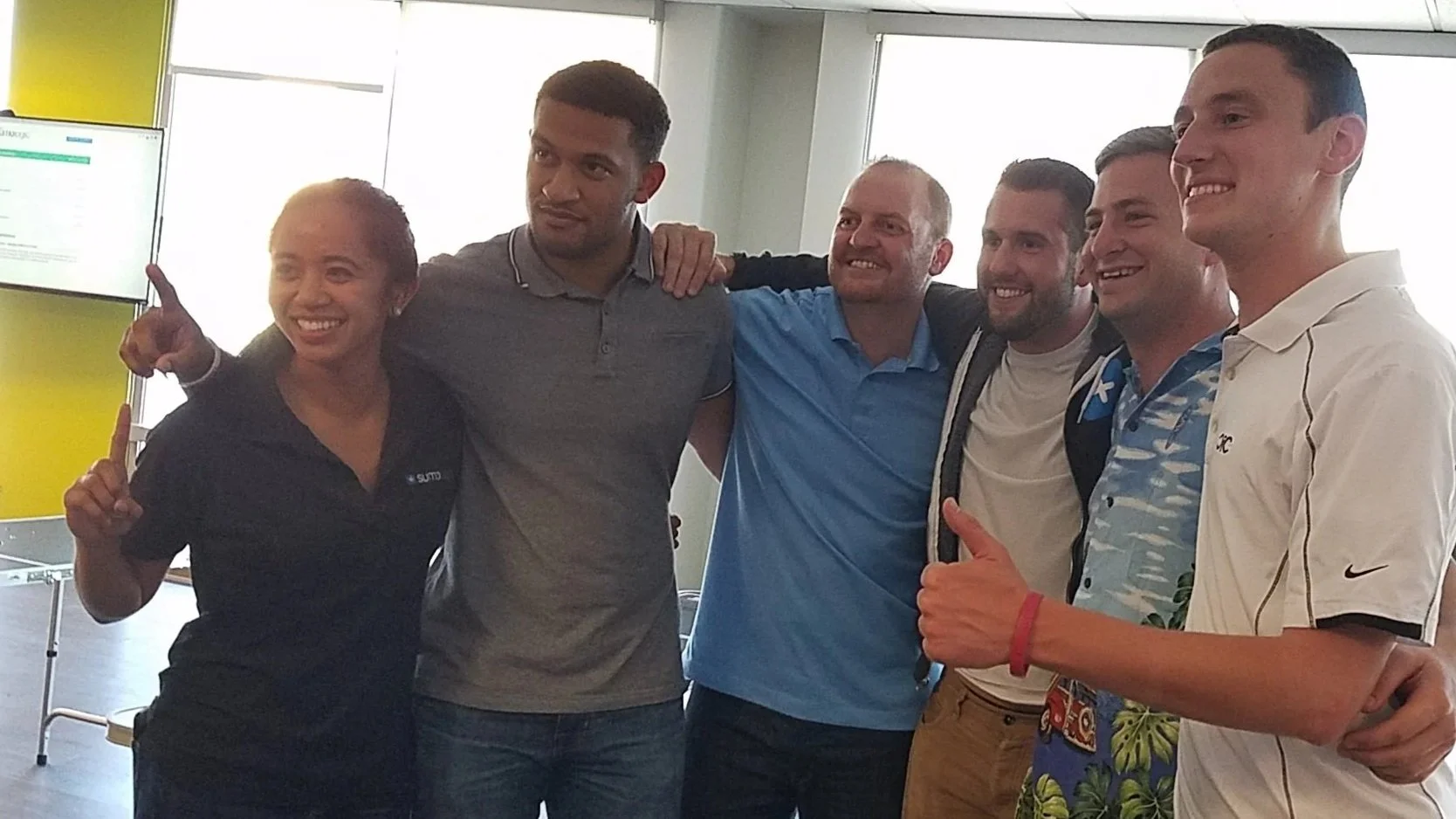 Group of six diverse people standing together indoors, smiling, with arms around each other, dressed casually.