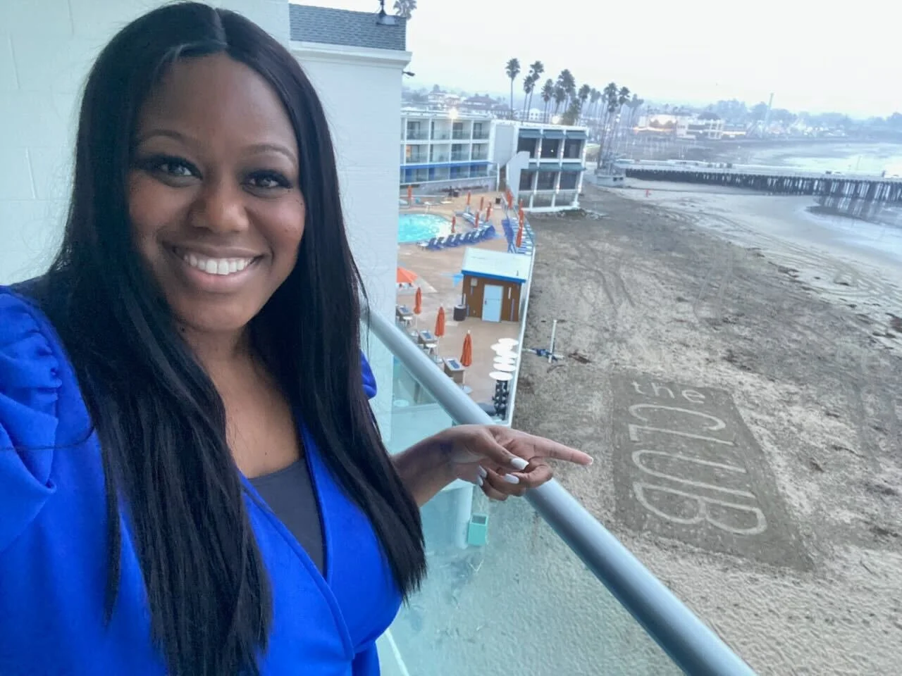 A woman in a blue jacket smiling and pointing at the sidewalk that reads 'the edge' near a beach with palm trees, buildings, and a pier in the background.