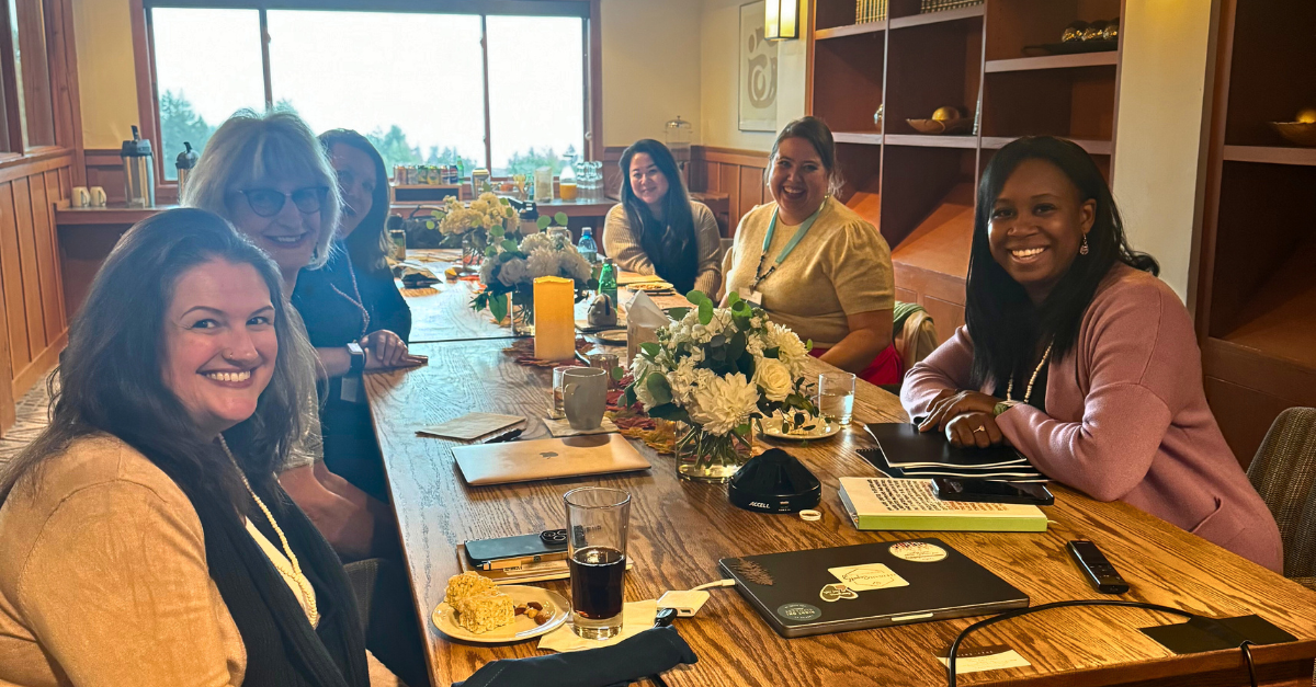 Group of women sitting around a conference table, smiling, with drinks and floral centerpieces.